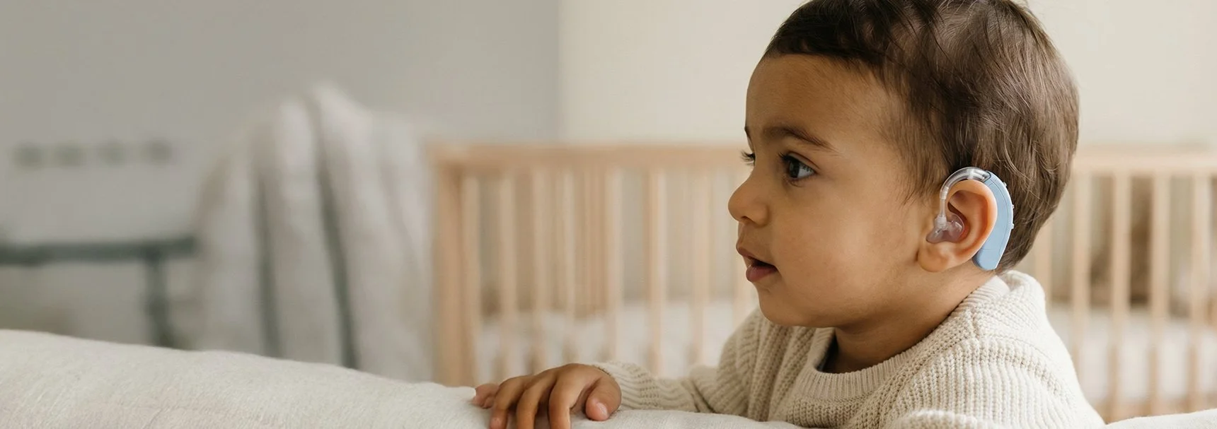 Young child with a hearing aid in his ear looking to the side in a nursery or bedroom.