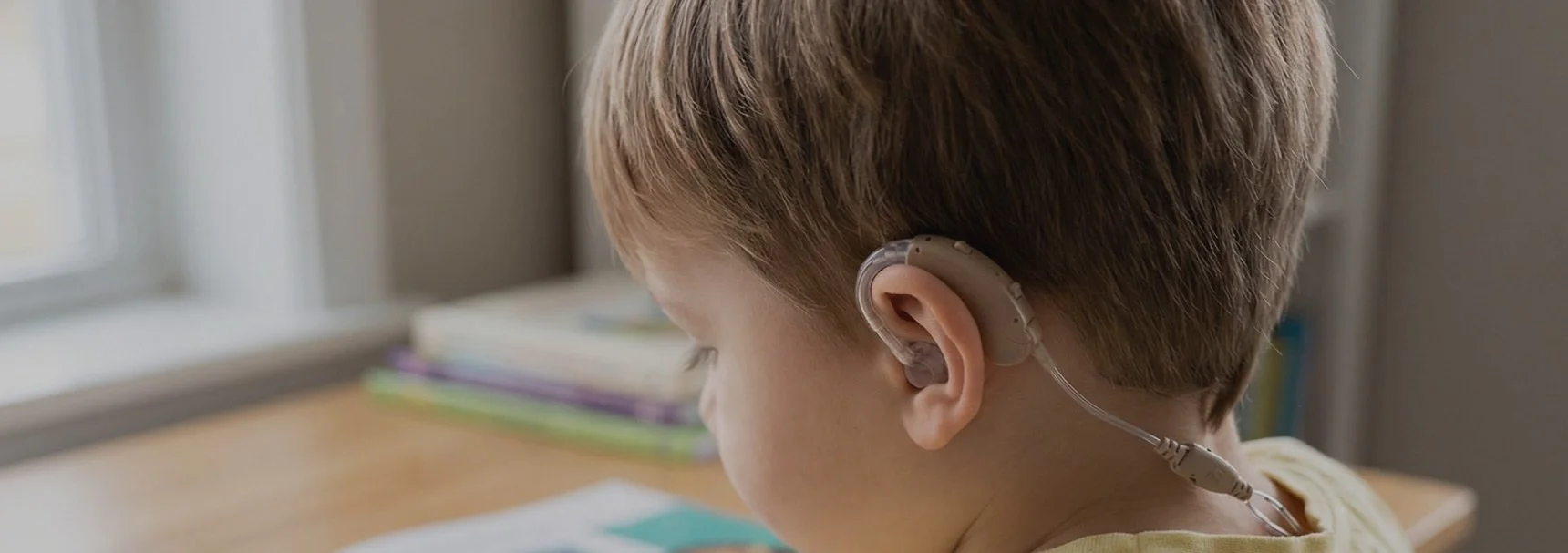 A young boy with light brown hair wearing a beige hearing aid in his right ear, sitting at a desk near a window, with a stack of colorful books in the background.