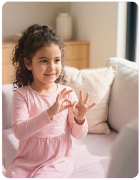Young girl with curly hair wearing a pink long-sleeve dress sitting on a beige couch, smiling and using hand gestures, in a living room with a wooden cabinet and a large window.
