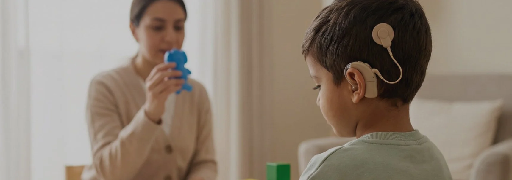 A boy sitting in a chair wearing a hearing aid and a woman holding a blue teddy bear-shaped object, possibly a toy or medical device, in a room with neutral-colored walls.
