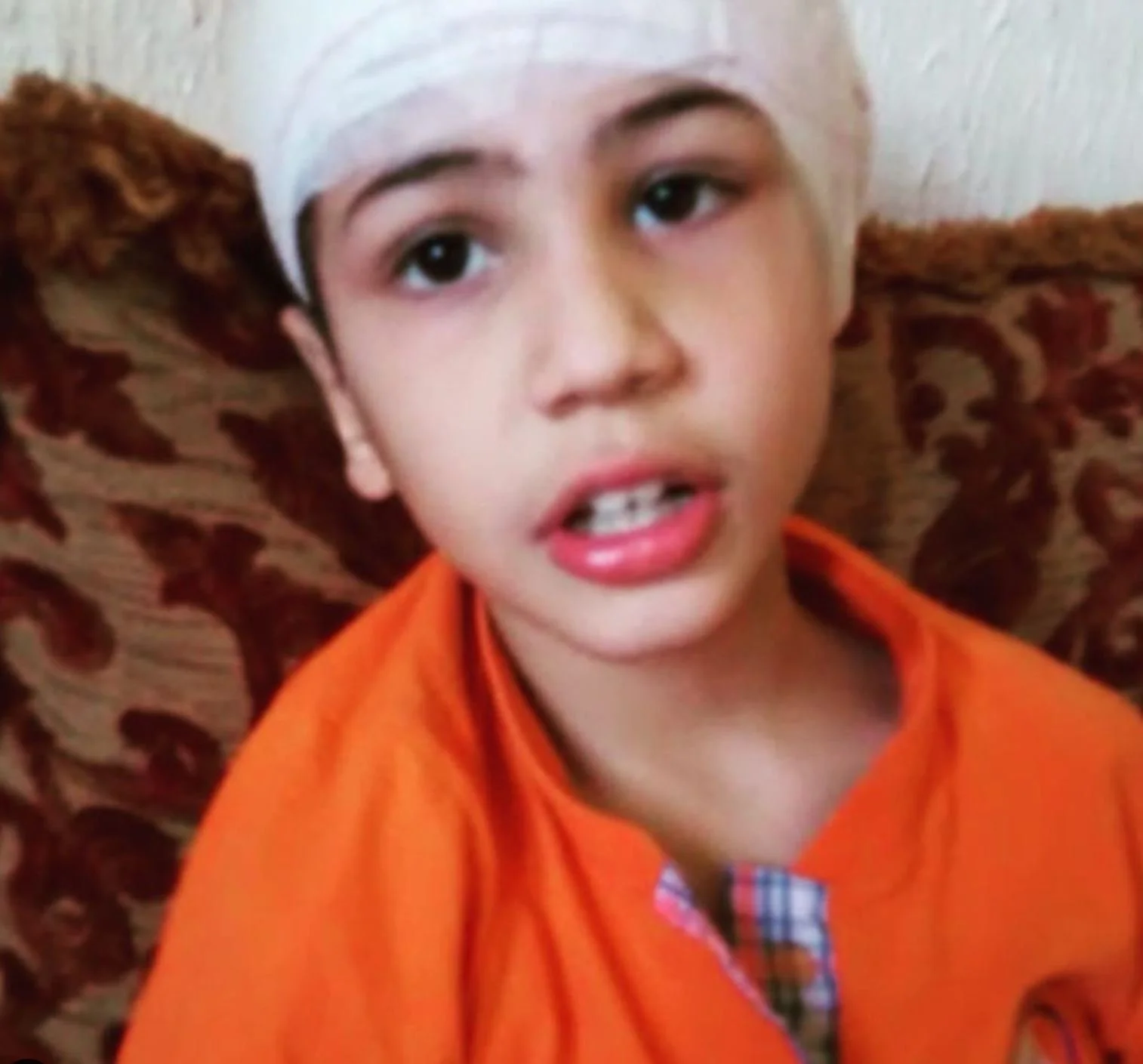 A young boy with a white headband, wearing an orange shirt, speaking or explaining something.