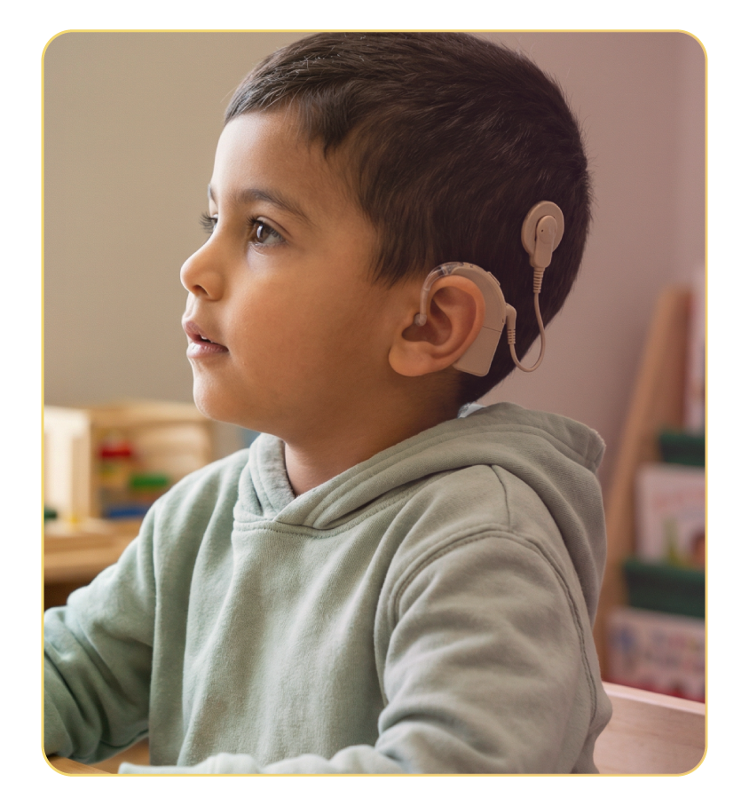 A young boy with short dark hair wearing a beige hoodie and a cochlear implant on his right ear, seated indoors with a neutral background.