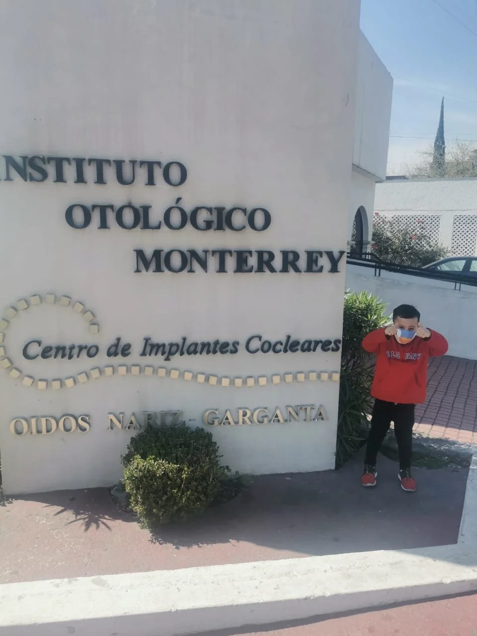 A young boy wearing a red hoodie and a face mask on a sunny day standing next to the sign for the Otolaryngology Institute of Monterrey, Mexico, with the words "Centro de Implantes Cocleares" and "Oidos Nariz Garganta" visible on the wall.