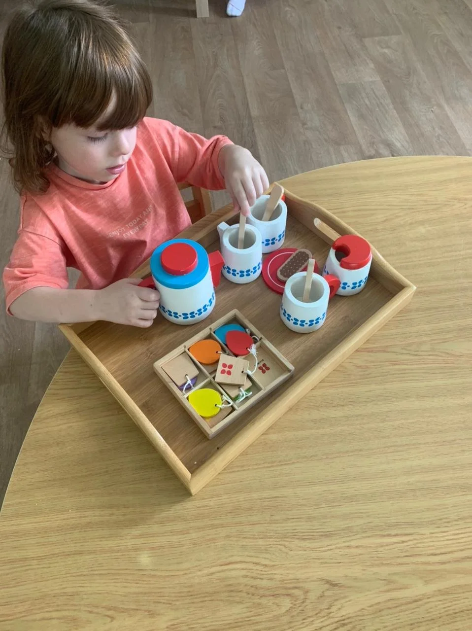A young girl playing with a toy set of pretend food and drink in a wooden tray.