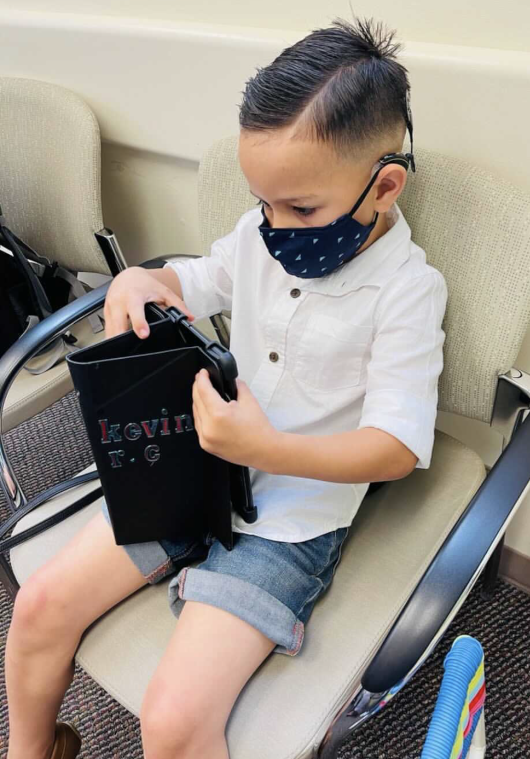 A young boy sitting on a beige chair, wearing a white shirt, denim shorts, a blue face mask, and looking at a black folder or binder.