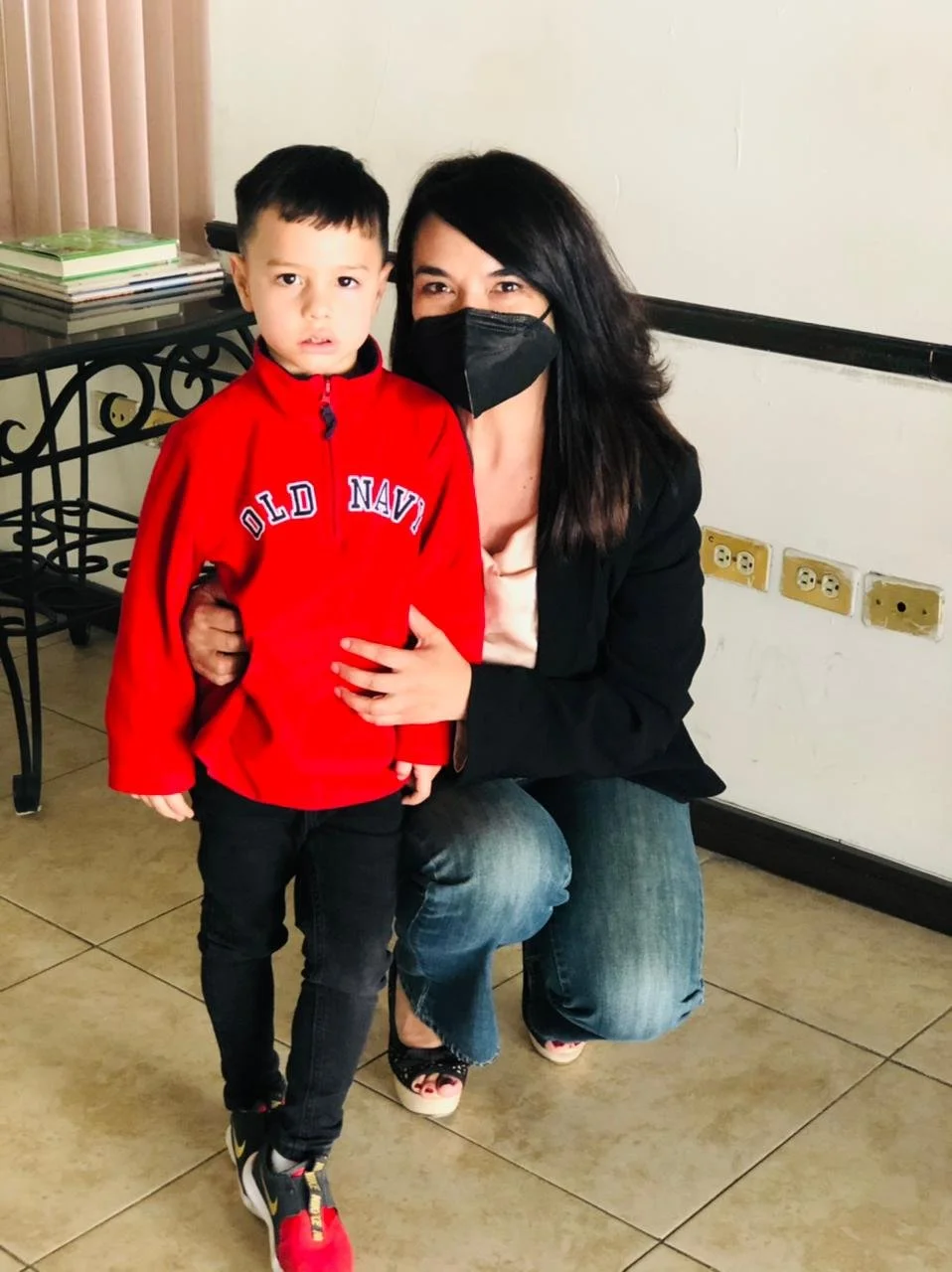 A woman with black hair wearing a black face mask and a dark jacket, sitting next to a young boy with short black hair wearing a red Old Navy jacket and black pants. They are indoors, with a table holding a stack of books and a wall with multiple ele