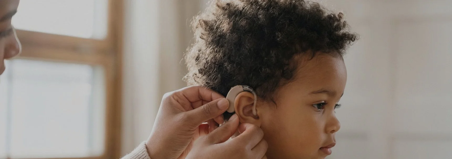 Close-up of a person adjusting a hearing aid in a young child's ear in a bright indoor setting.