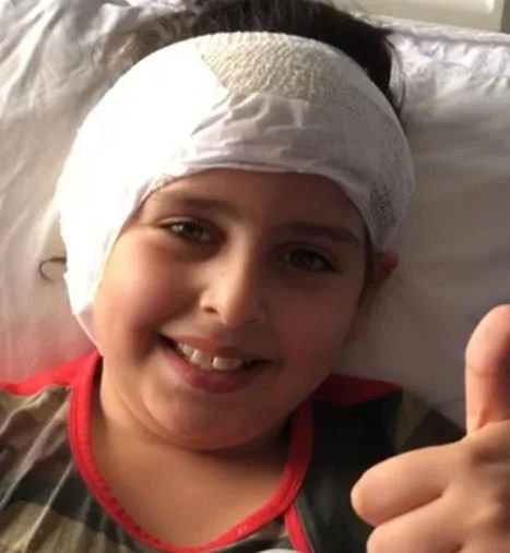 Young boy smiling and giving a thumbs-up, with a bandaged head, lying in bed.