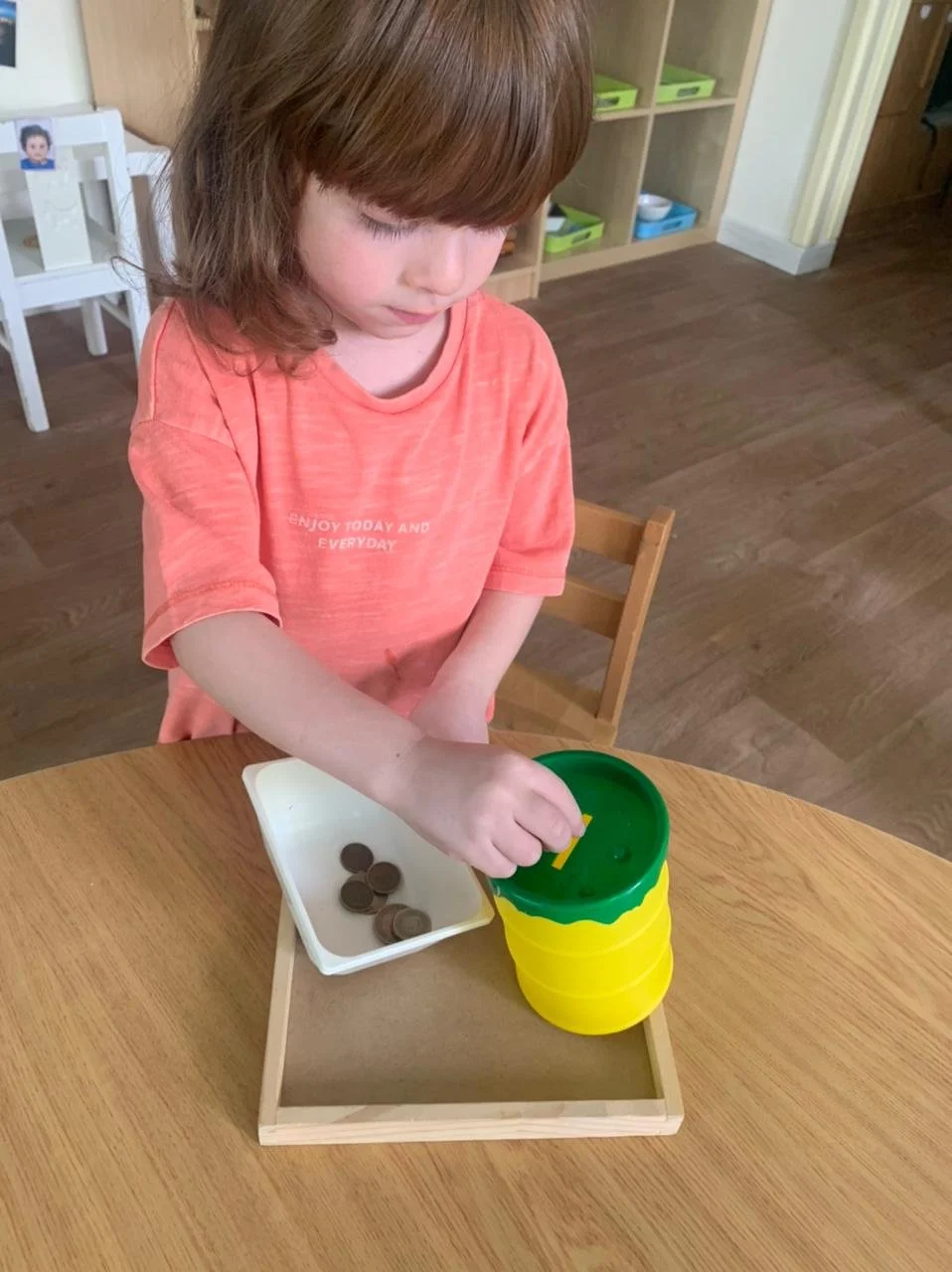A young girl with light skin and brown hair, wearing an orange t-shirt, is engaging with a toy that resembles a stacking game on a wooden table. The toy has a yellow base and a green top. A small white tray with coins is nearby.