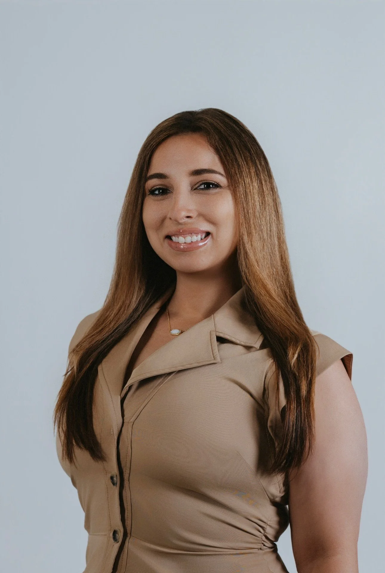 Portrait of a woman with long brown hair, wearing a beige sleeveless dress and a necklace, smiling against a plain light gray background.