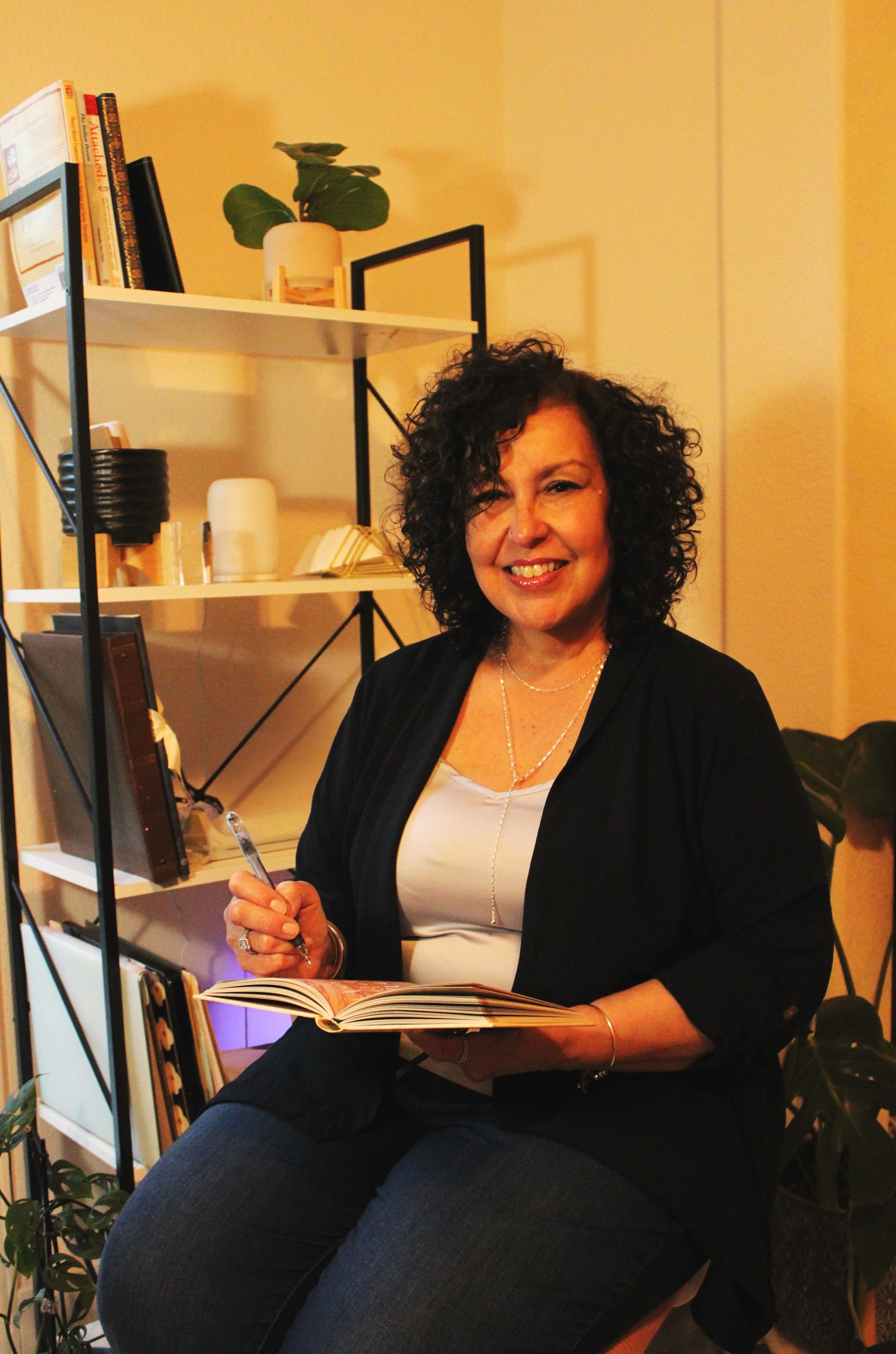 Meet Connie- Parent coach with curly dark hair smiling while sitting at a table, holding a pen over an open notebook ready to work collaboratively to support you navigating your child's challenging behaviors.