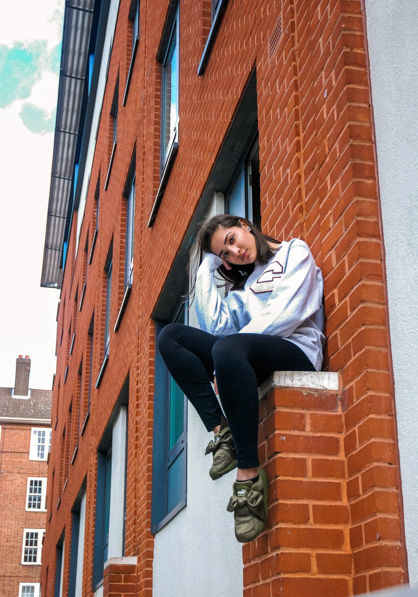 Young woman with dark hair, wearing a white jacket, black leggings, and green sneakers, sitting on a brick ledge outside a red brick building with blue window frames, looking at the camera.