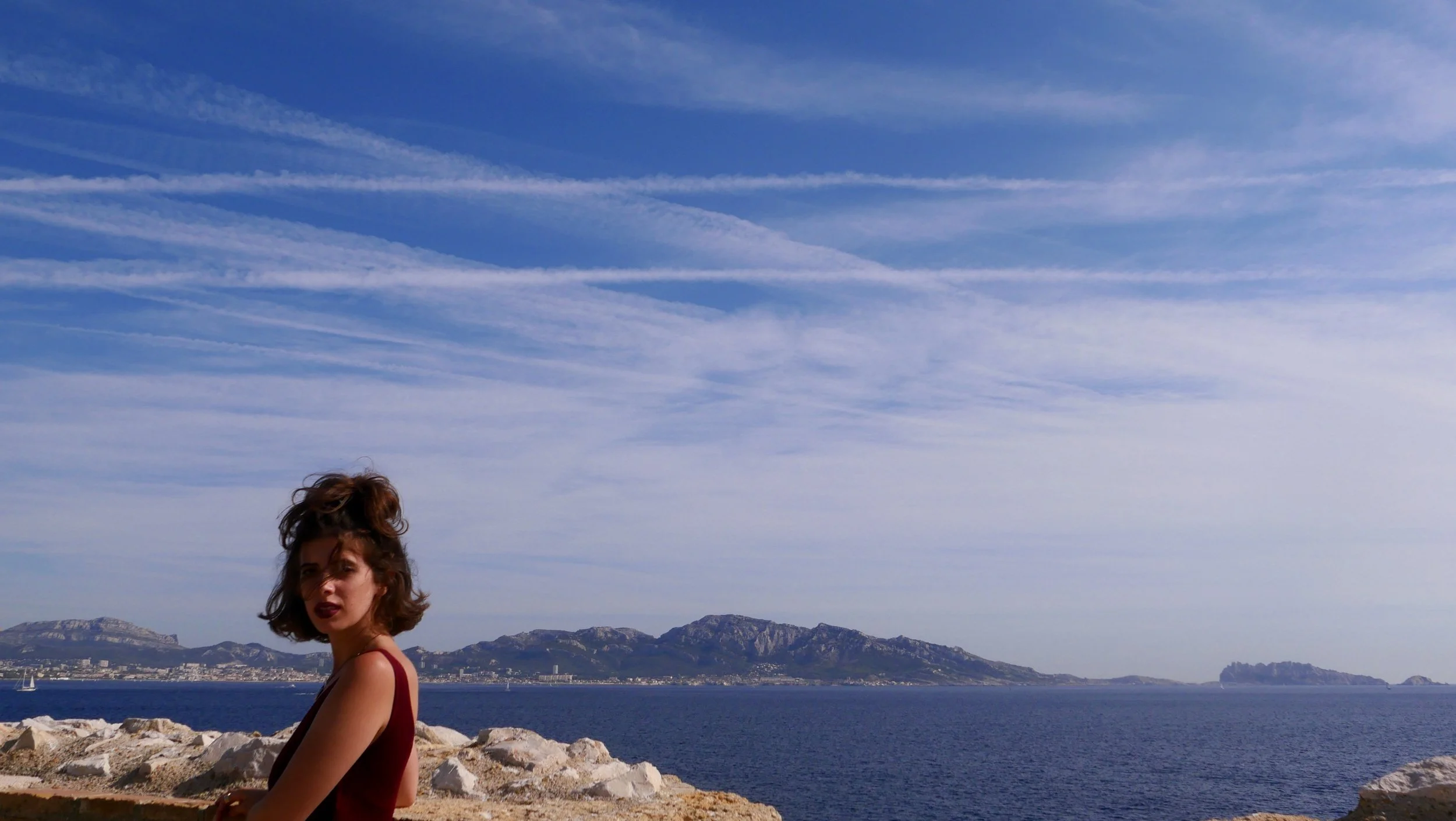 A young woman with curly hair and wearing a sleeveless top, sitting on rocks near a body of water under a partly cloudy sky, with mountains in the distance.