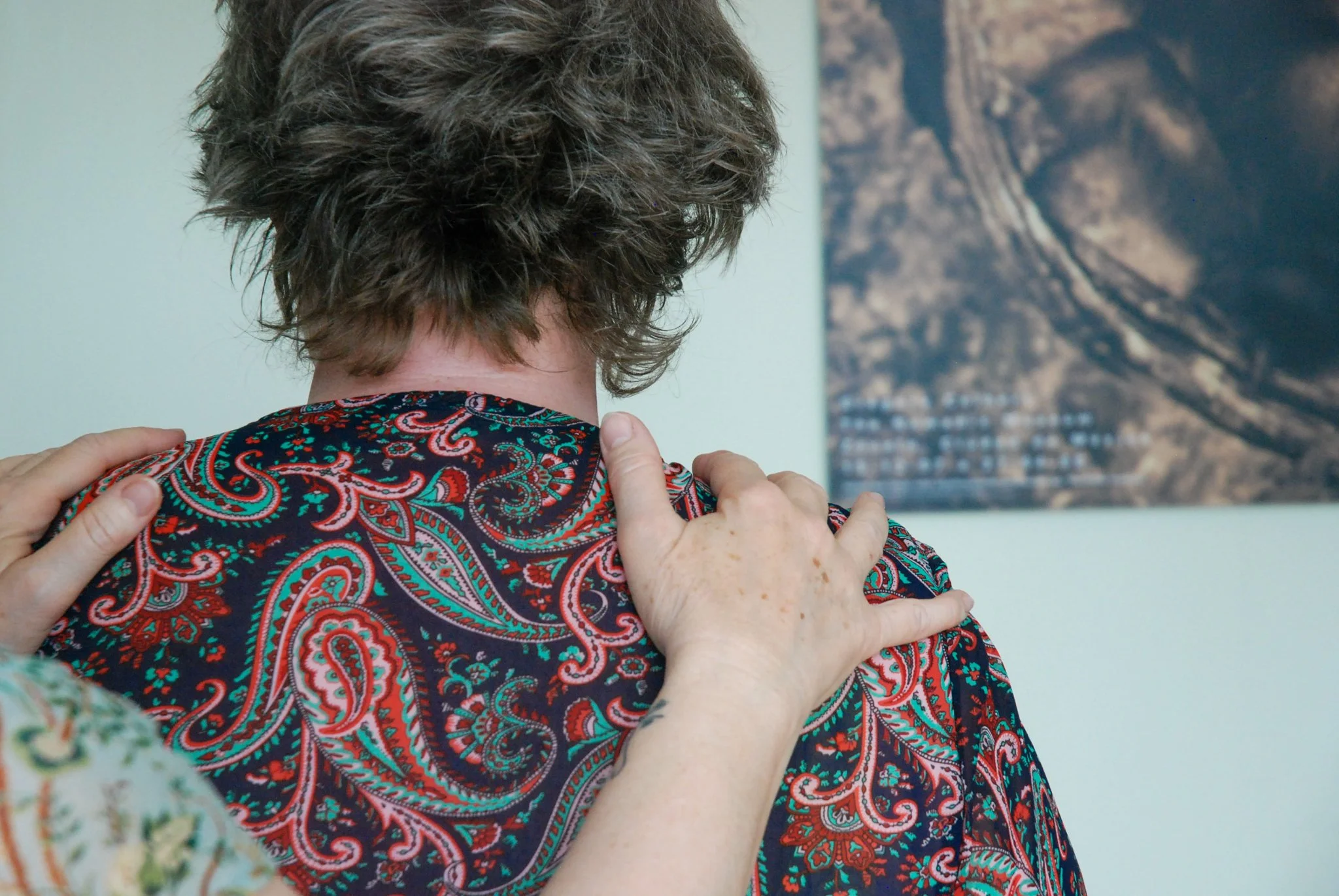 A person with short, curly gray hair wearing a colorful paisley-patterned shirt and another person with light green patterned clothing touching their shoulders, standing in front of a wall with a mountain landscape painting.