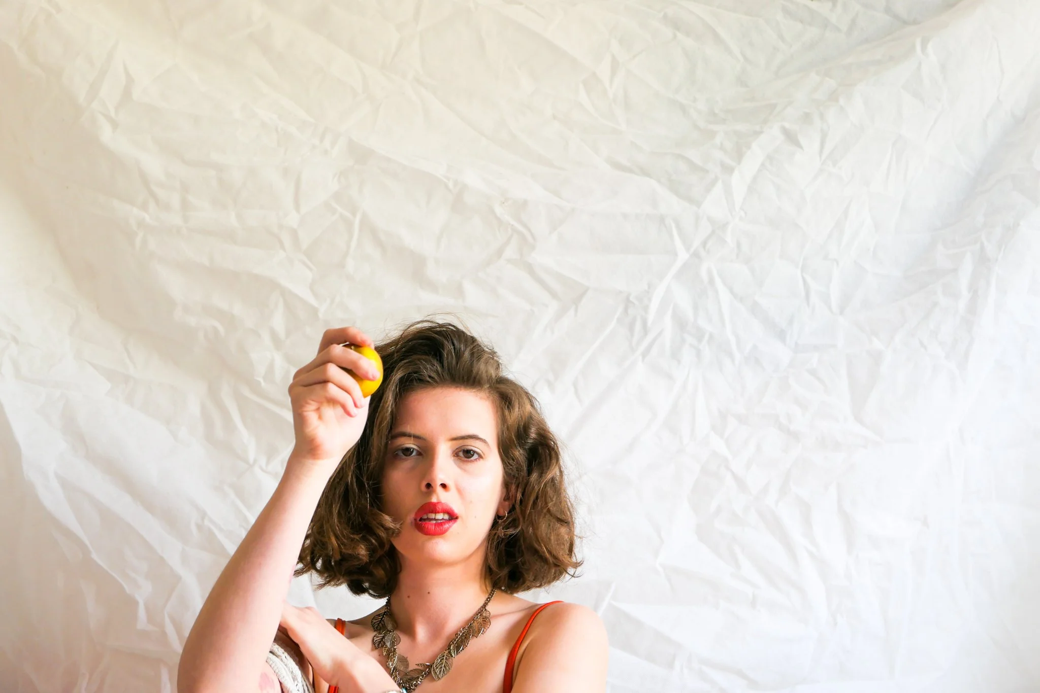A woman with curly brown hair and red lipstick holding a yellow object above her head, standing against crumpled white paper background.