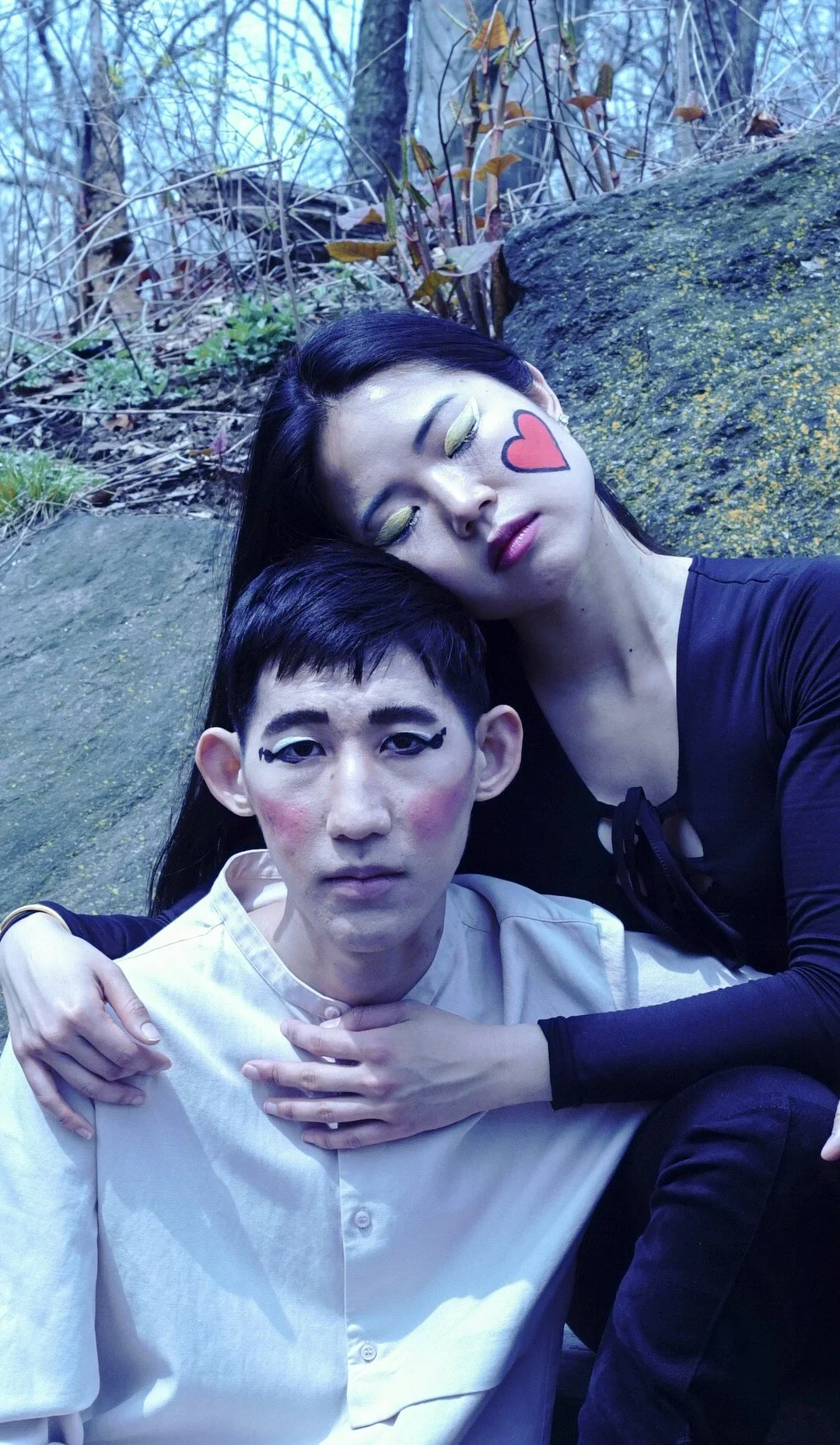 A person with artistic makeup sitting outdoors, with a woman with closed eyes and painted lips, leaning her head on their shoulder in a natural setting with rocks and trees.