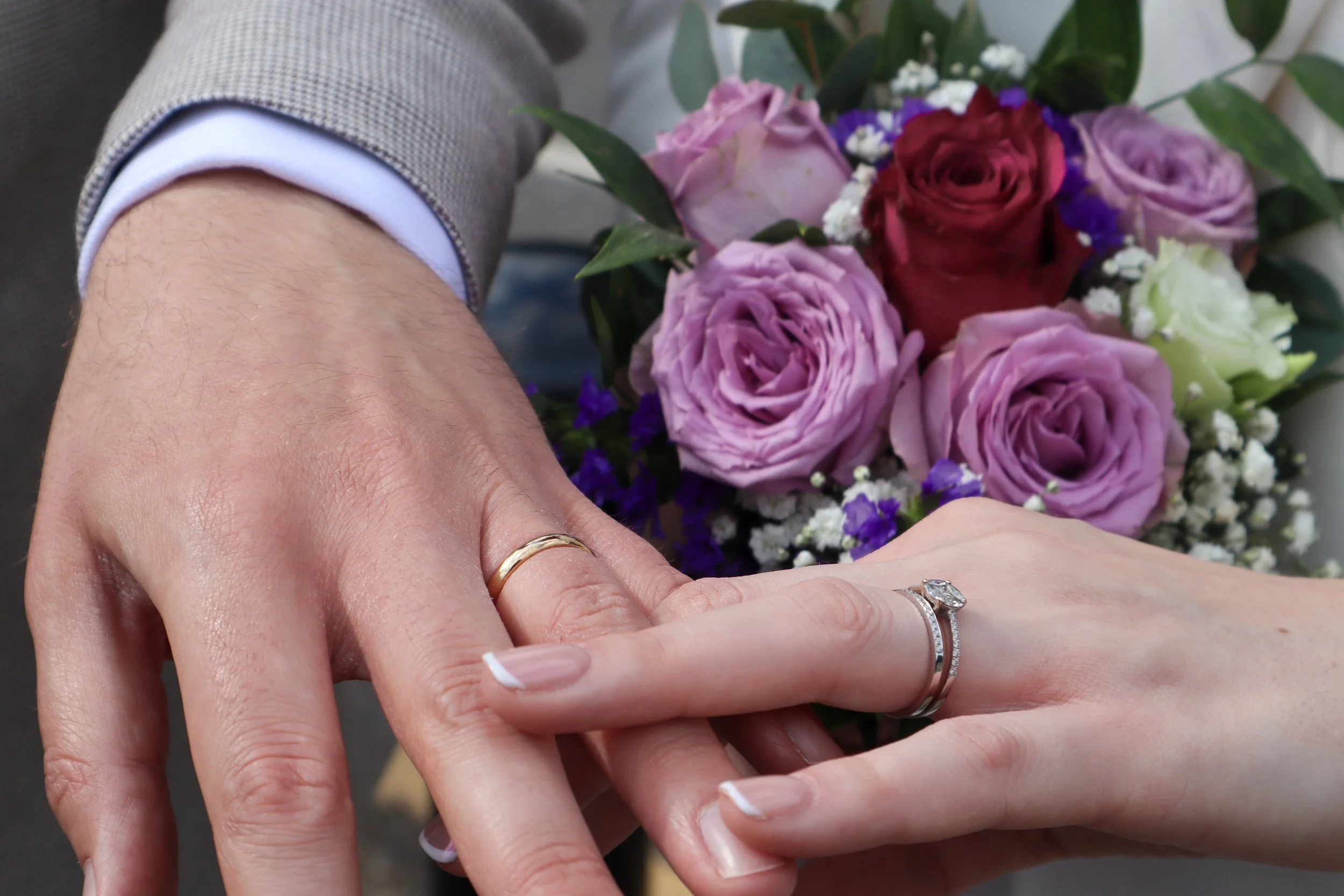 Close-up of two hands with wedding rings, in front of a bouquet of pink, purple, and white roses, with leaves and small white filler flowers.