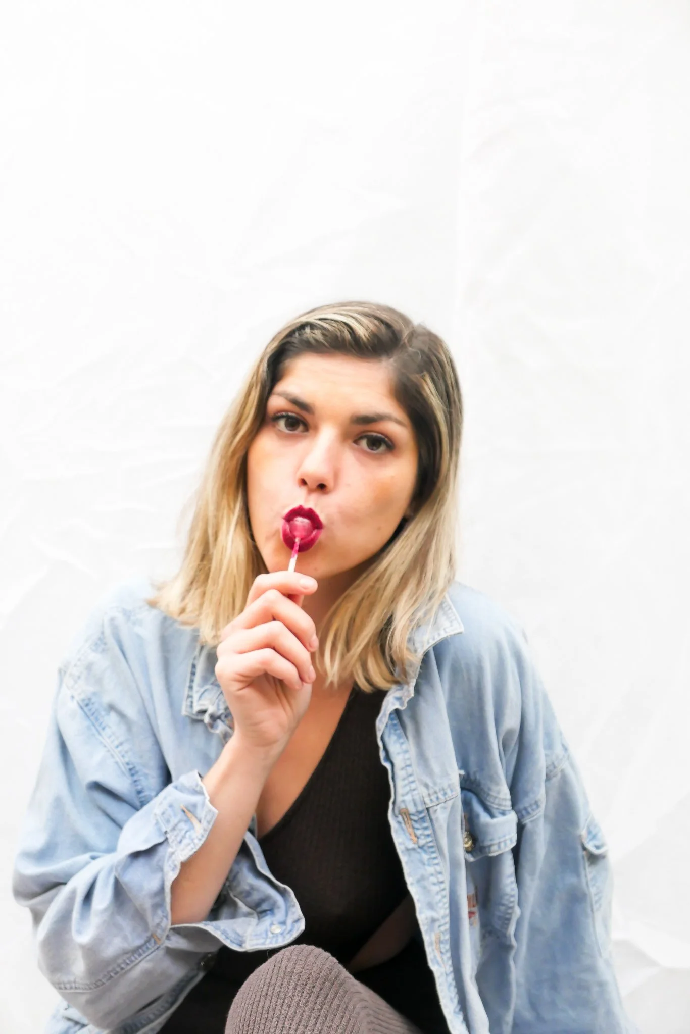Young woman with blonde hair, wearing a black top and light denim jacket, sitting against a plain white background, holding a red lollipop near her lips, with a neutral expression.