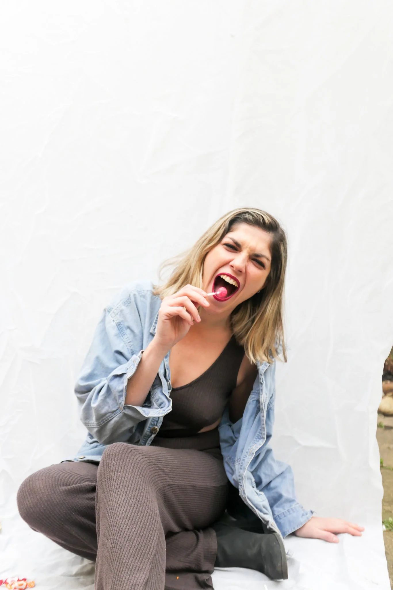 A young woman with blonde hair, wearing a black top and a blue denim jacket, is sitting cross-legged outdoors on a white surface, holding a pink lollipop near her mouth and showing a playful expression.