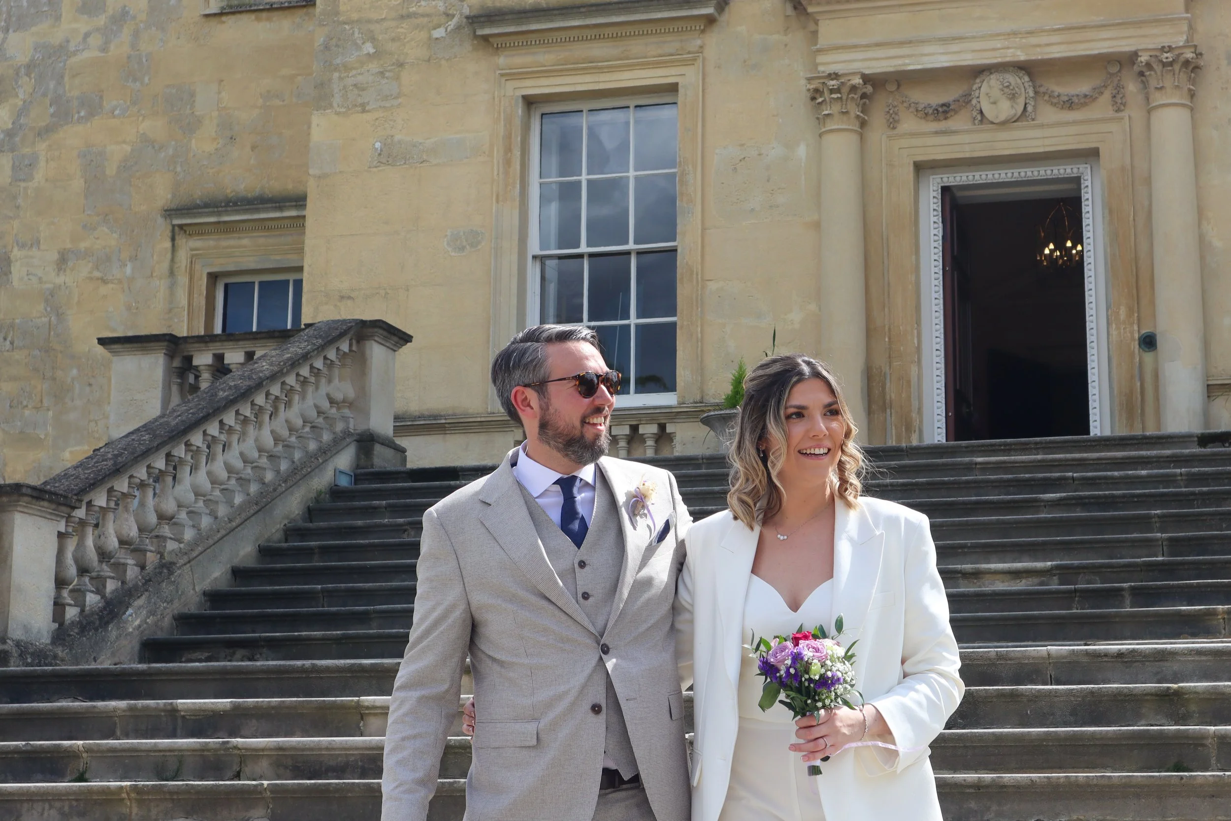 A bride and groom standing on stone steps outside a historic building, the groom wearing a light gray suit with sunglasses, and the bride in a white outfit holding a bouquet, both smiling.
