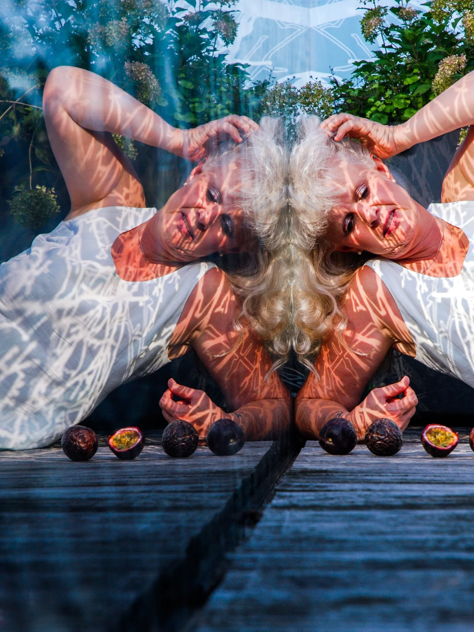 Two elderly women with white hair lying on their backs on a wooden surface, surrounded by plants and flowers, with multiple halved passion fruits lined up in front of them, as they smile and enjoy the moment.