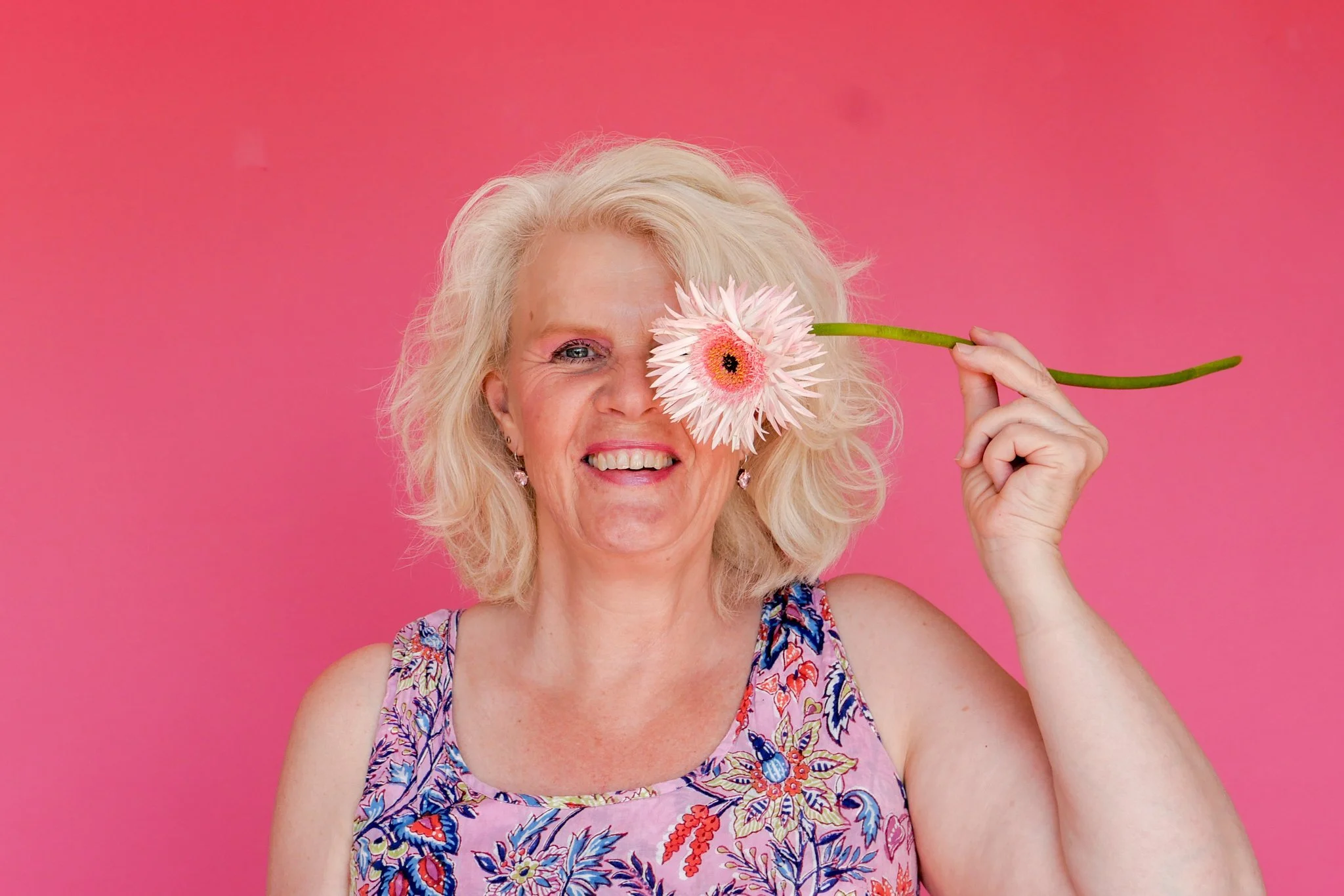 A woman with blonde curly hair holding a pink and white flower over her eye, smiling against a pink background.