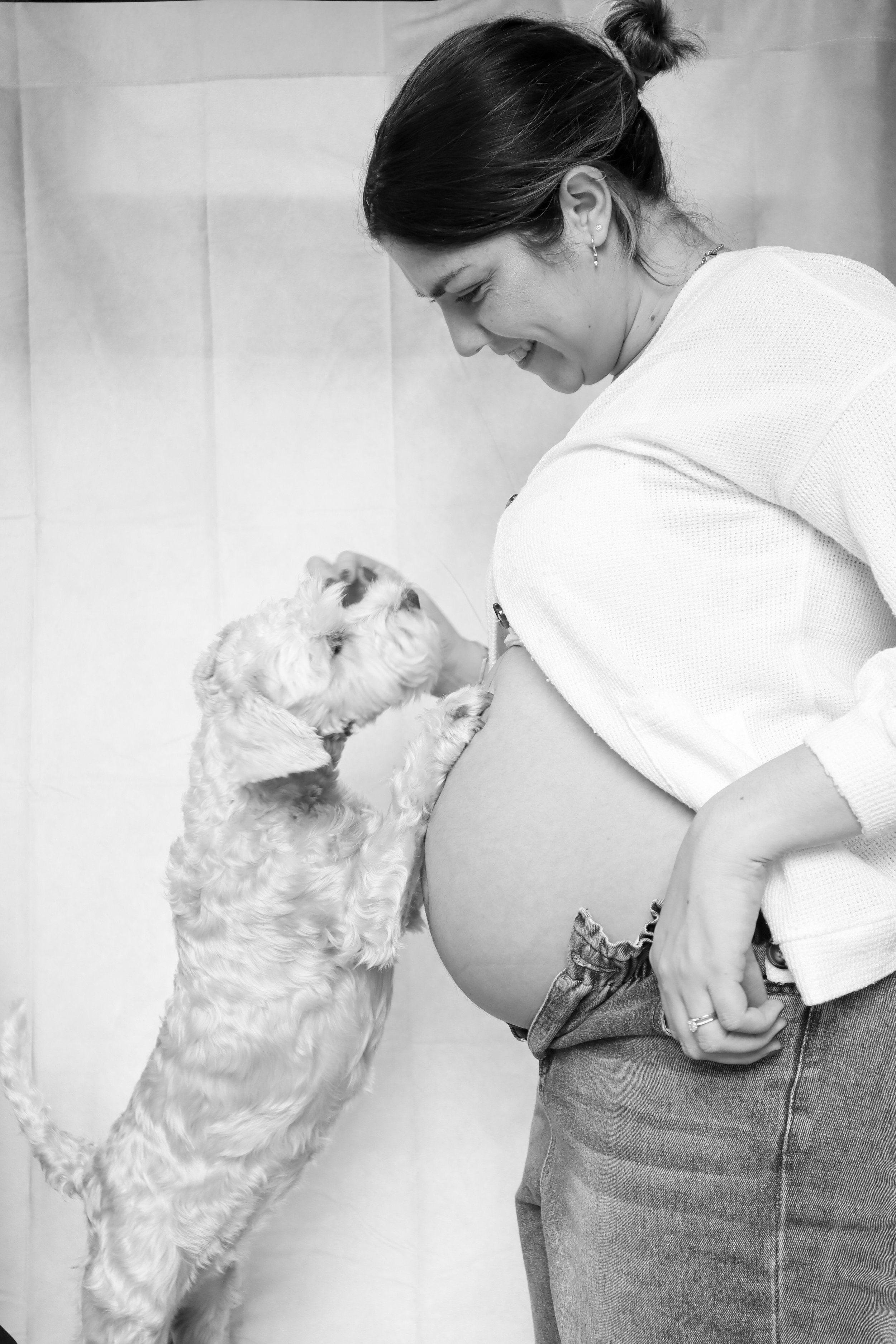 A woman with dark hair in a bun smiling while looking down at a small dog. The dog is standing on its hind legs with its front paws on her tummy, looking up at her. The woman is wearing earrings, a white shirt, and jeans with a belt. The background i