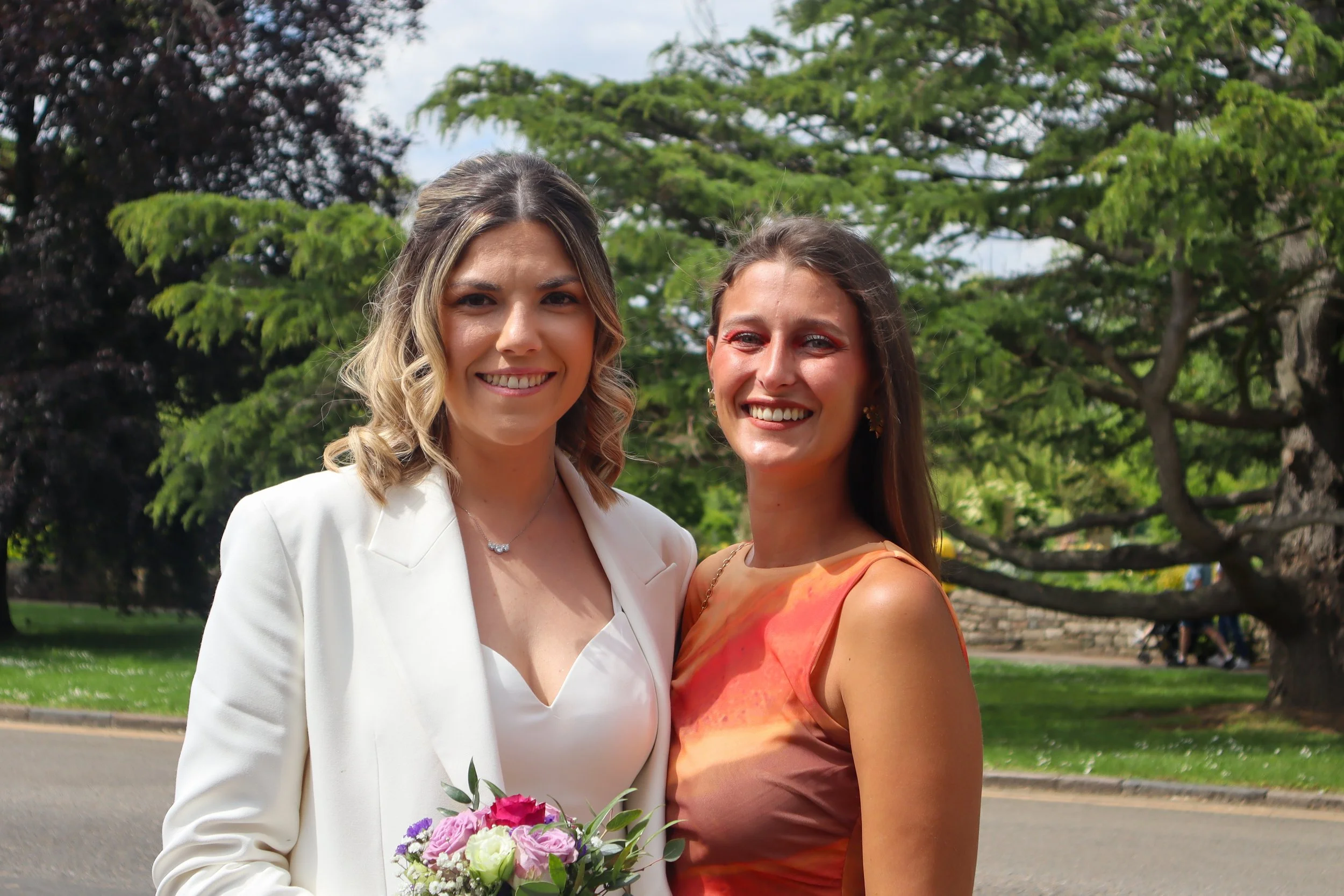 Two smiling women, one in a white blazer holding a bouquet of pink and white flowers, standing outdoors in a park with green trees in the background.