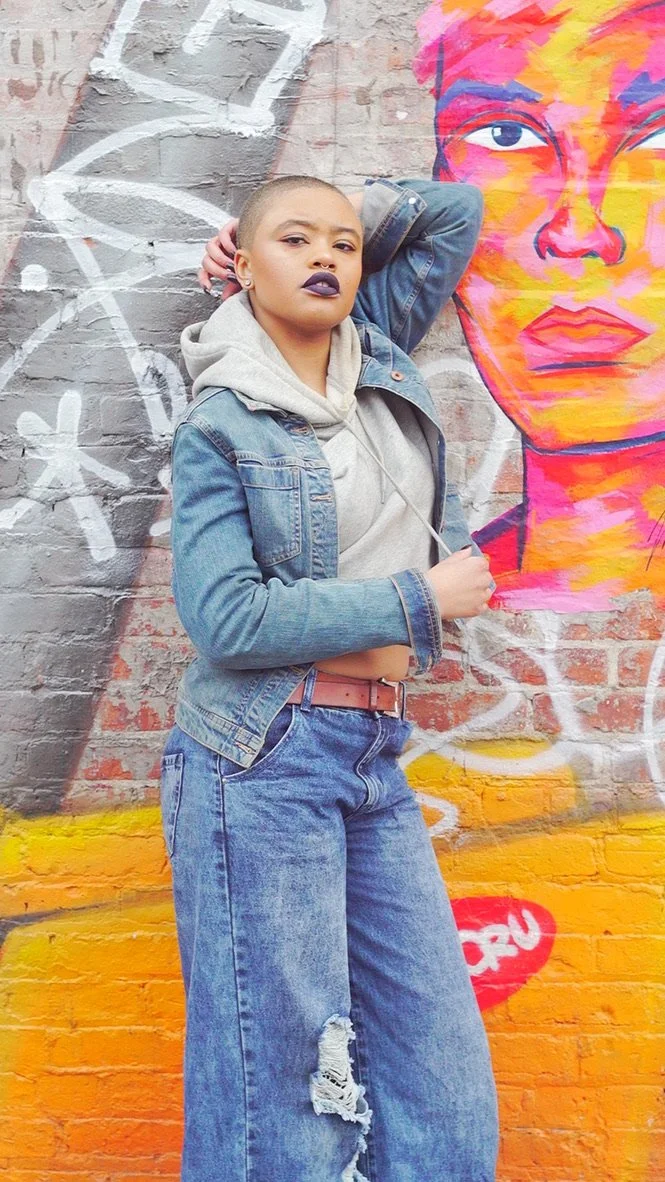 A person with short hair, wearing a denim jacket, gray hoodie, and ripped jeans, standing in front of a colorful mural of a woman's face on a brick wall.