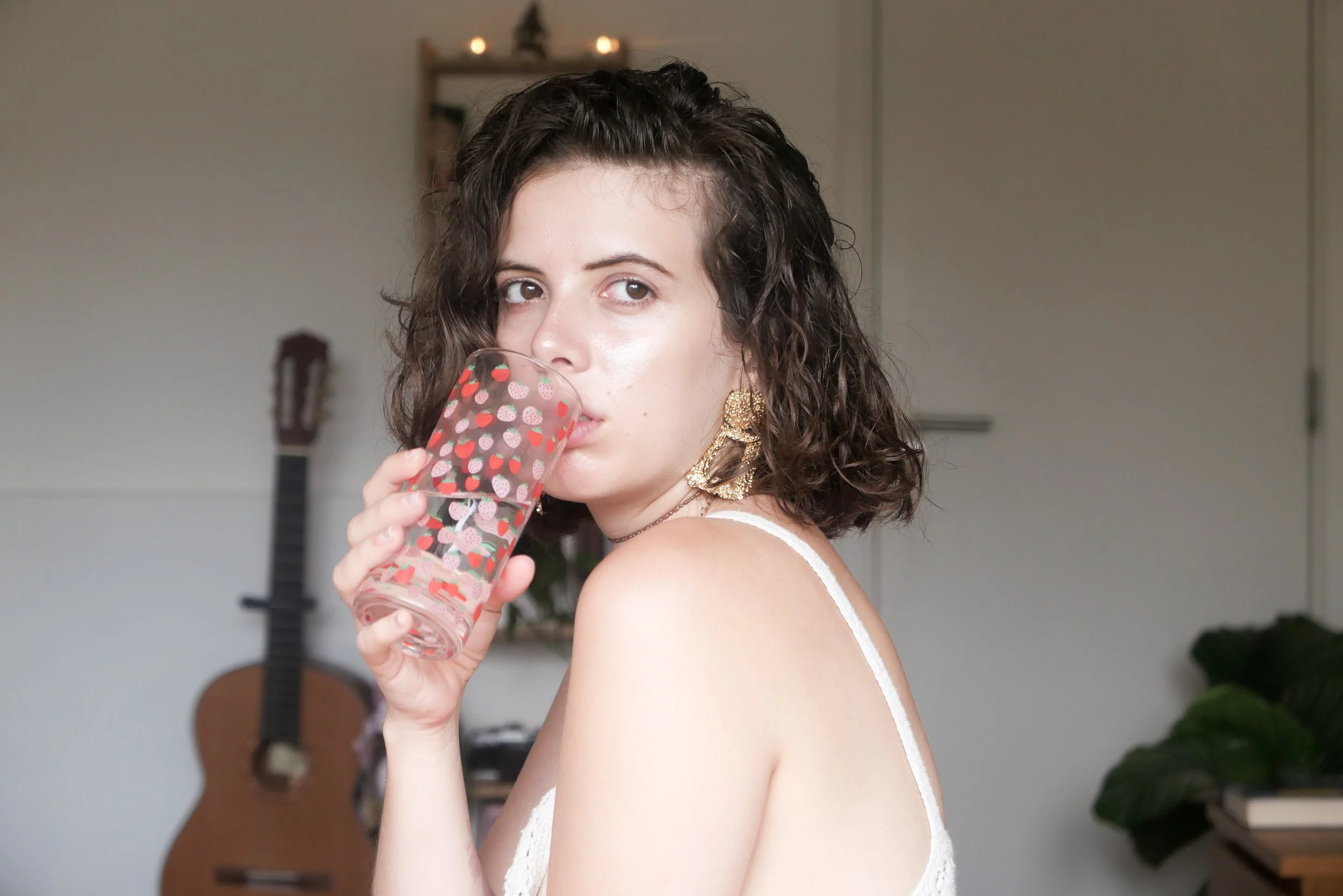 A woman with curly dark hair and gold earrings drinking from a glass with a strawberry pattern, in a room with a guitar and some plants in the background.