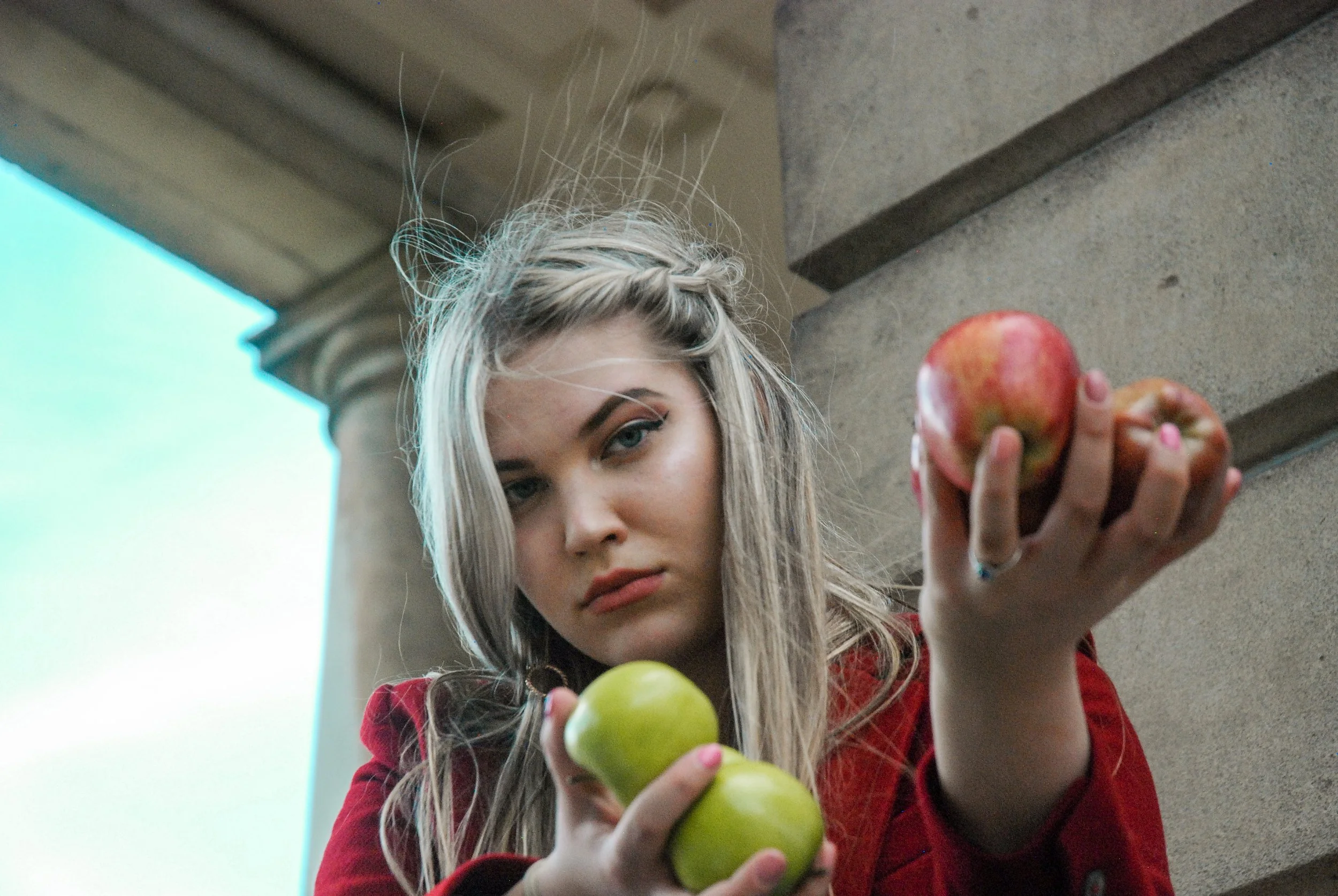 Young woman with blonde hair and light skin holding green and red apples, looking down at the camera, with a stone architectural structure in the background.