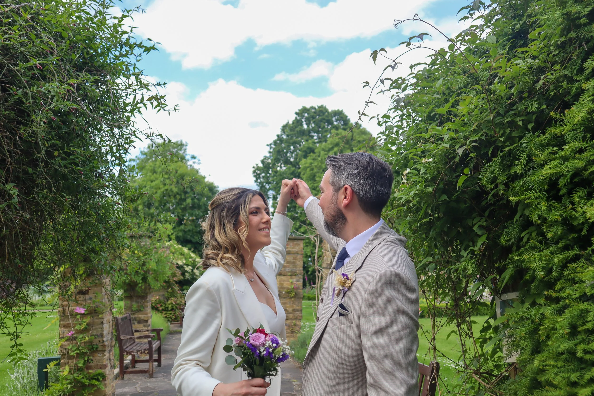 A couple stands close together outdoors in a lush, green garden with trees and plants, sharing a dance or moment. The woman, with long wavy hair, holds a bouquet of purple flowers, and the man, with short gray hair and a beard, is dressed in a gray s