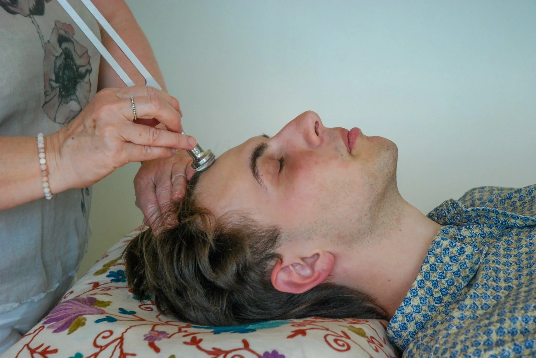 A person lying down with eyes closed, receiving a cosmetic treatment with a small metallic device on their forehead. The person has brown hair and is wearing a patterned shirt, while the practitioner has hands with a ring and a beaded bracelet, using