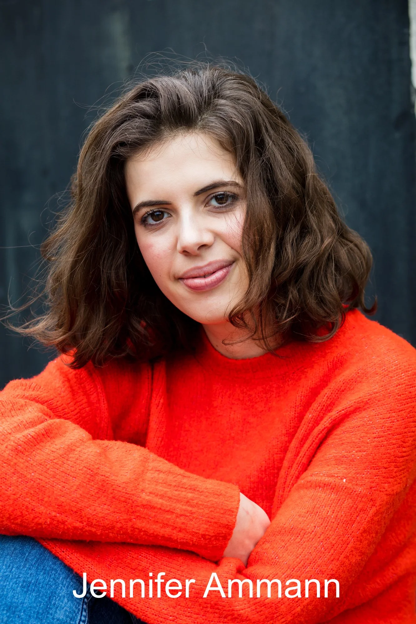 Close-up portrait of a woman with brown wavy hair and brown eyes wearing a bright orange sweater, smiling softly, with a dark background.