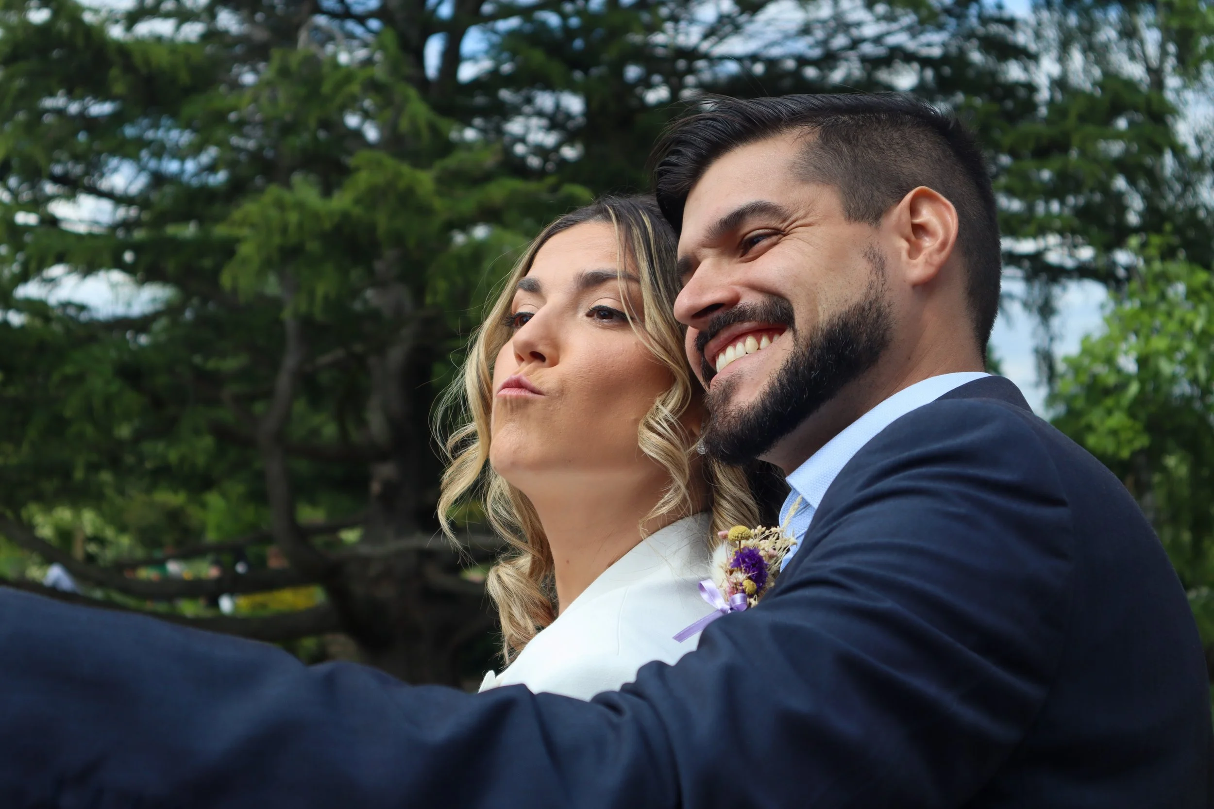A smiling man and woman taking a selfie outdoors with trees in the background, dressed in formal attire, likely at a wedding.