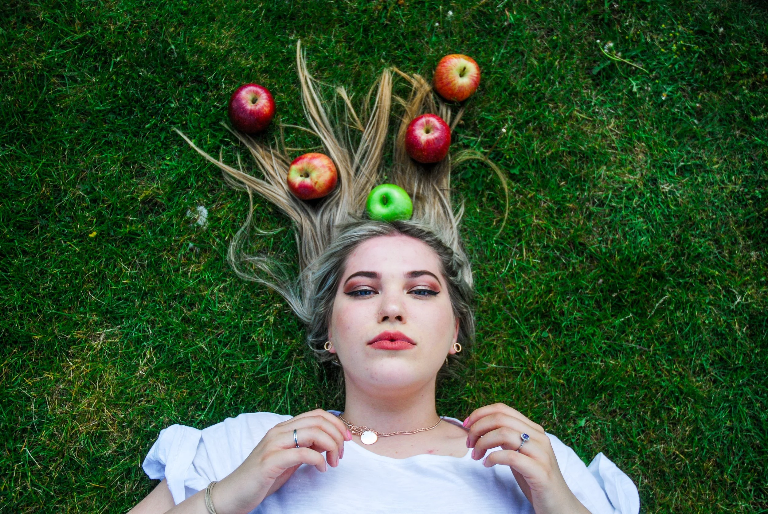 A young woman with blonde hair lying on green grass, holding jewelry, with several red and one green apple arranged in her hair.