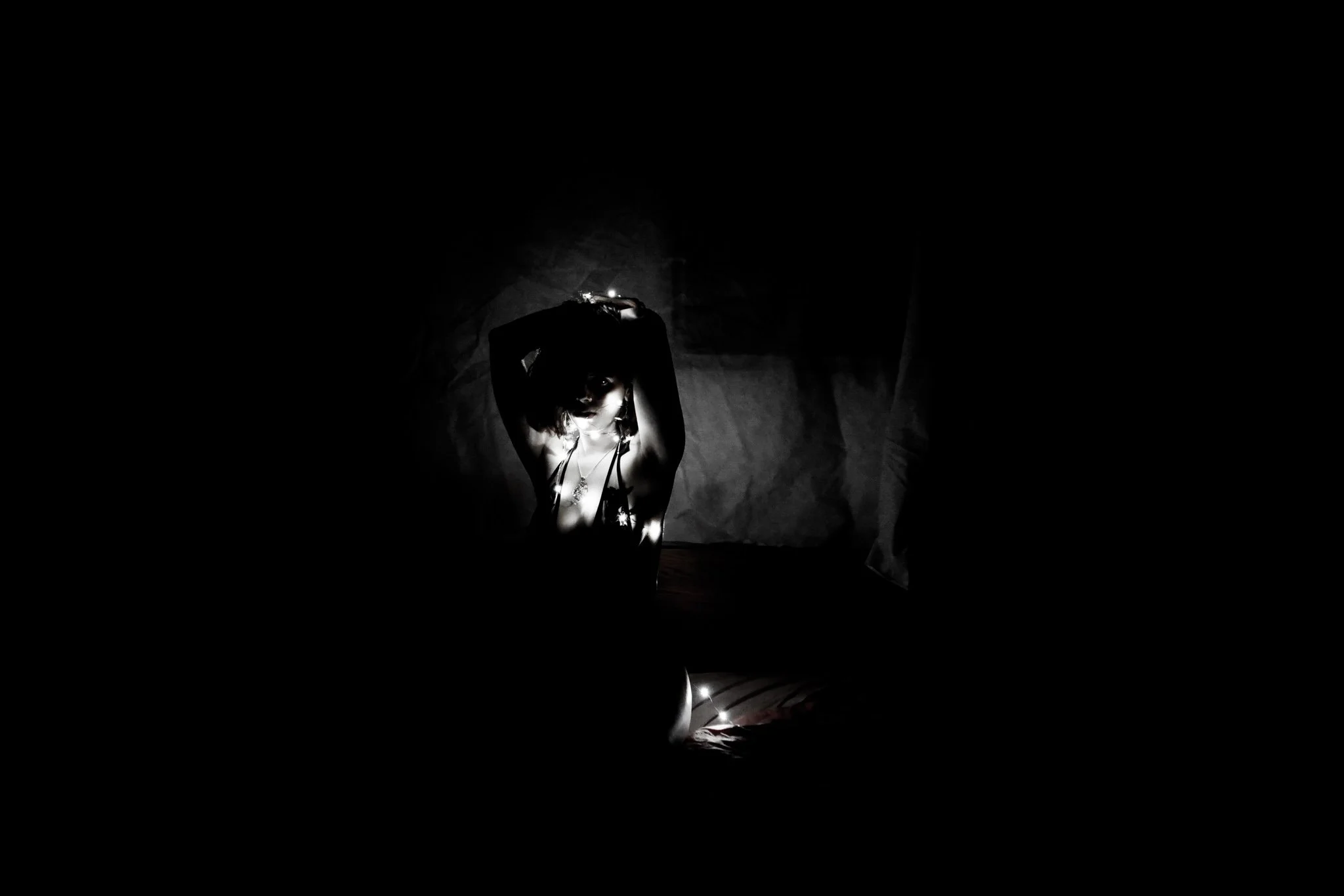 A woman with dark hair and makeup posing in a dark setting with dramatic lighting, her arms raised above her head, wearing a necklace and revealing clothing.
