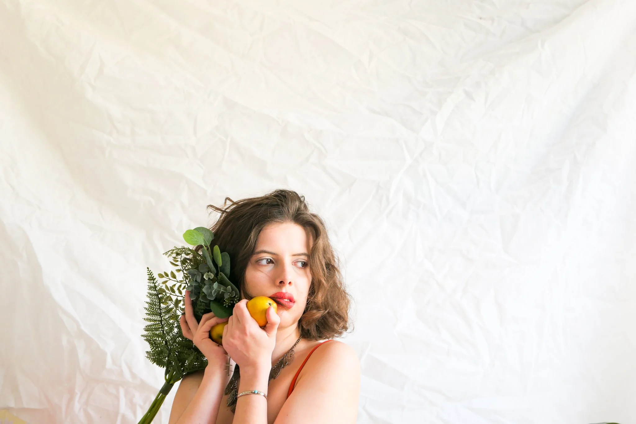 A woman with wavy brown hair holding a bouquet of flowers and fruits, including a yellow apple, near her face against a plain, wrinkled white background.