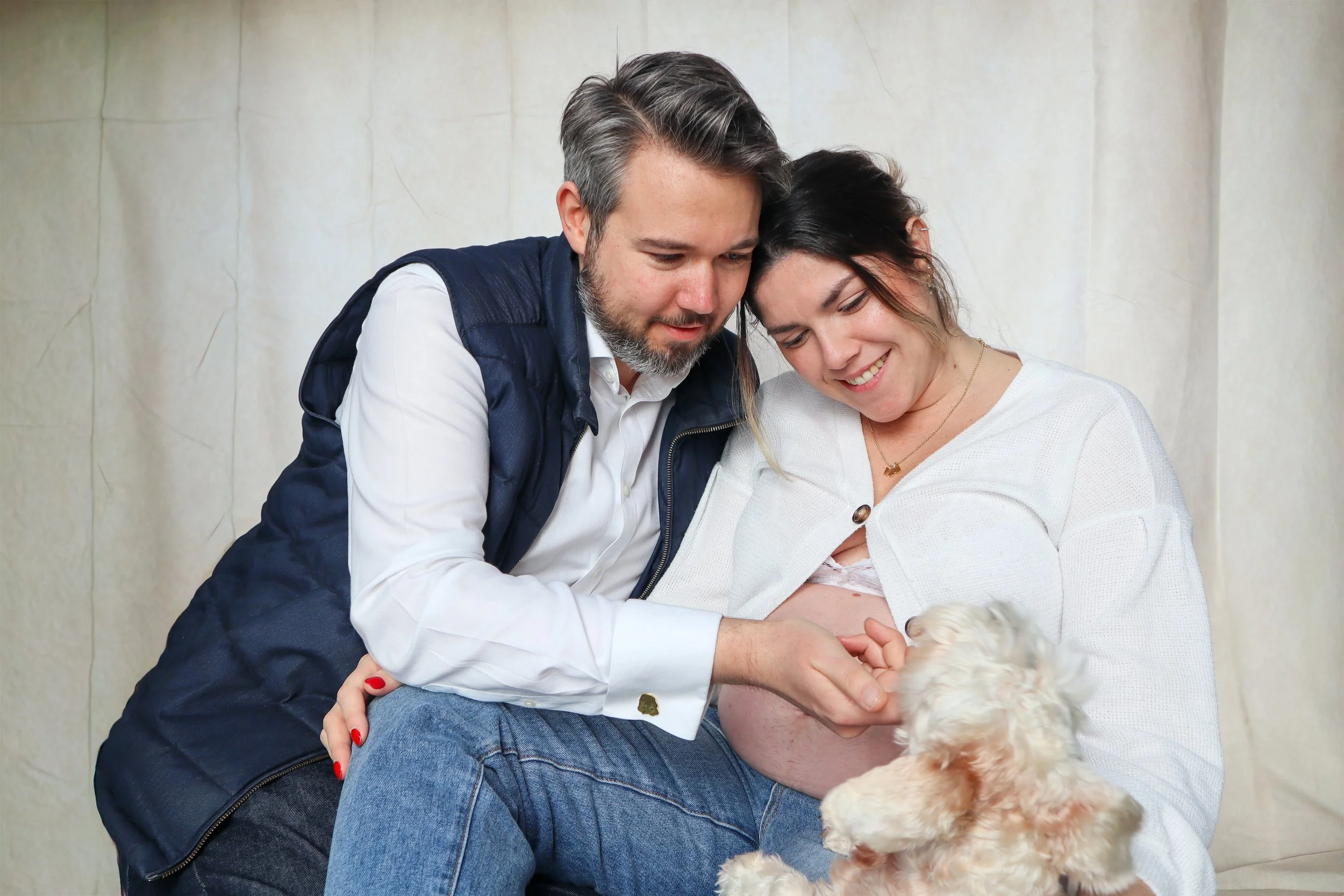 A couple cuddling and smiling while holding a small puppy, with a plain light-colored background.