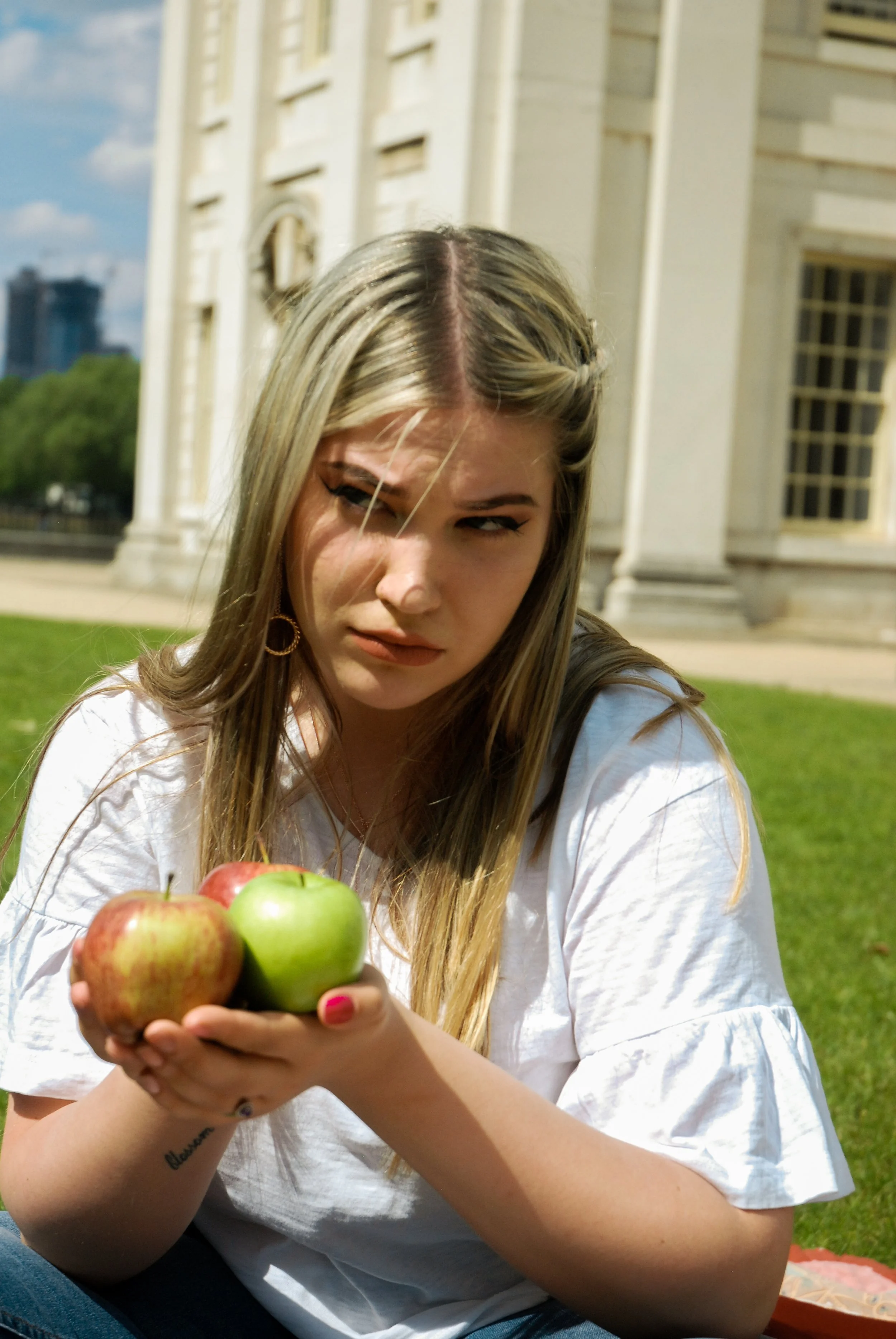 A young woman with long blonde hair is sitting outdoors in front of a large historical building, holding a small bunch of apples in her hand, with a contemplative or puzzled expression.
