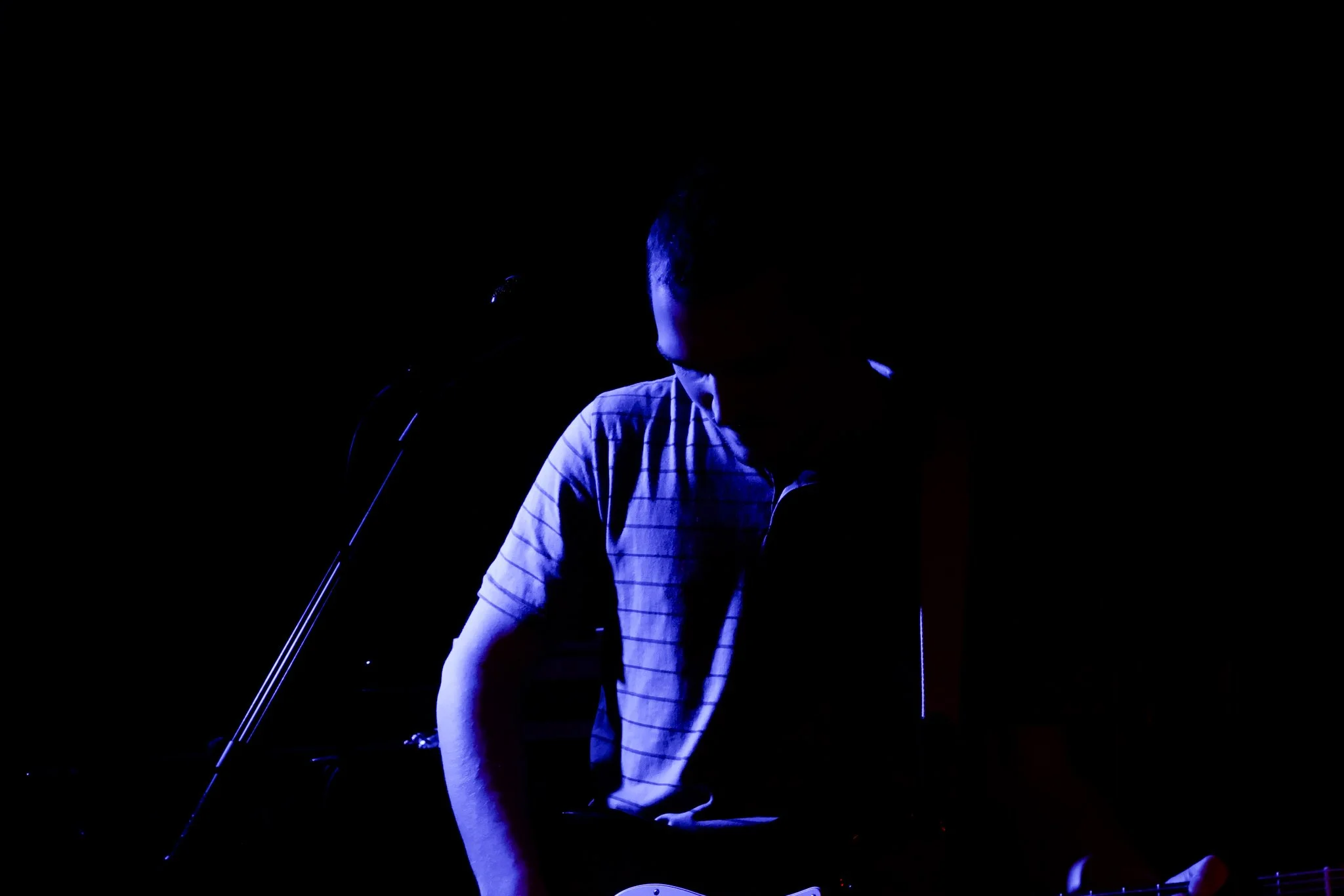 Person on stage, partially illuminated by blue light, standing in front of microphone, wearing a striped shirt.