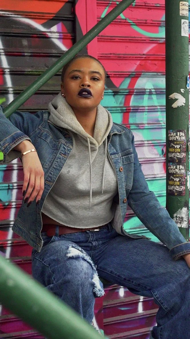 A woman with short hair, dark lipstick, and septum piercing, wearing a gray hoodie and denim jacket, sitting on a step in front of a graffiti-covered metal door, with a green pole and graffiti stickers nearby.