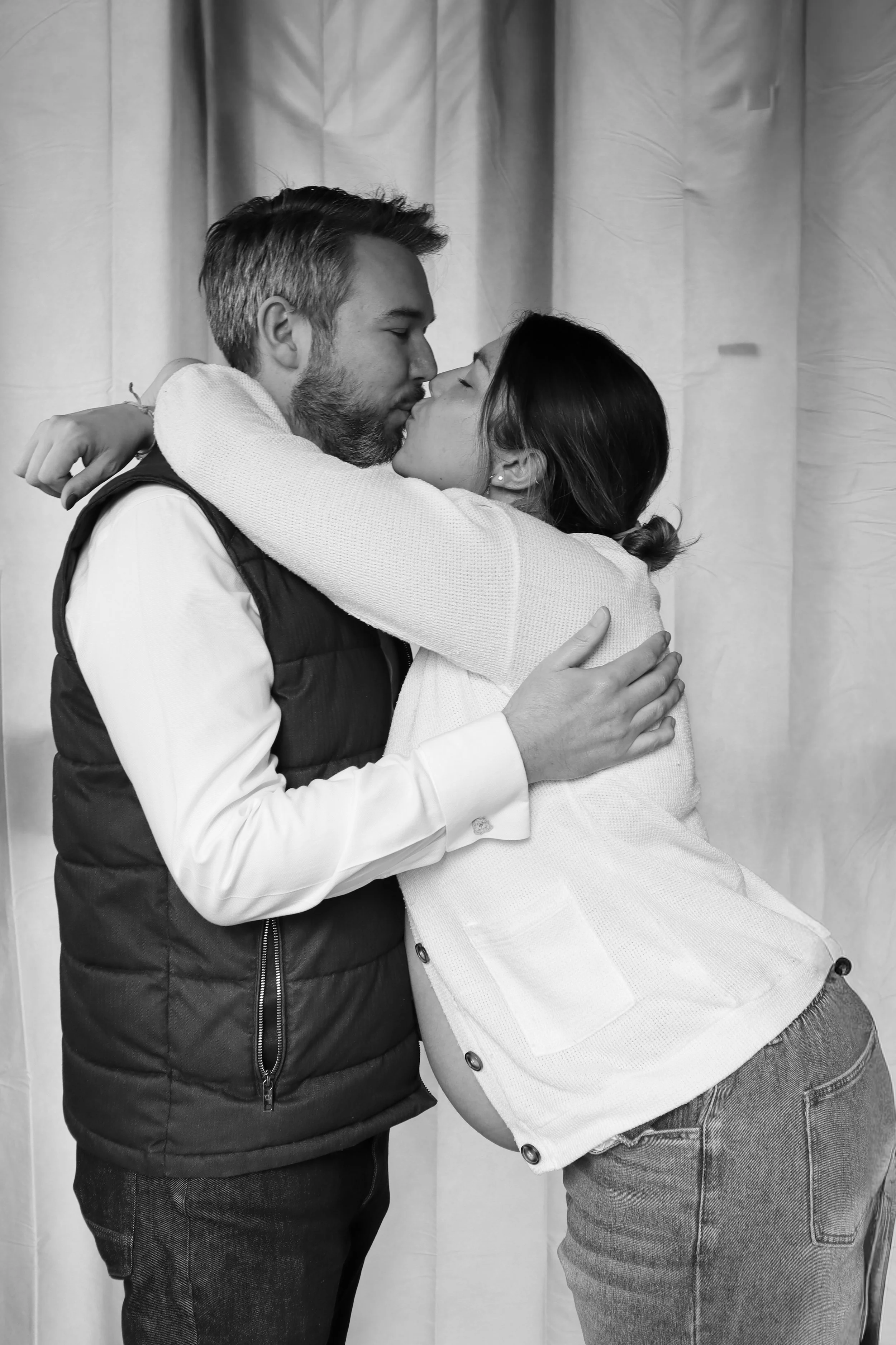 A man and woman are embracing tightly, about to kiss, with their eyes closed. The man is wearing a collared shirt and a puffy vest, and the woman is wearing a white jacket and jeans. They are standing in front of a plain, light-colored background.