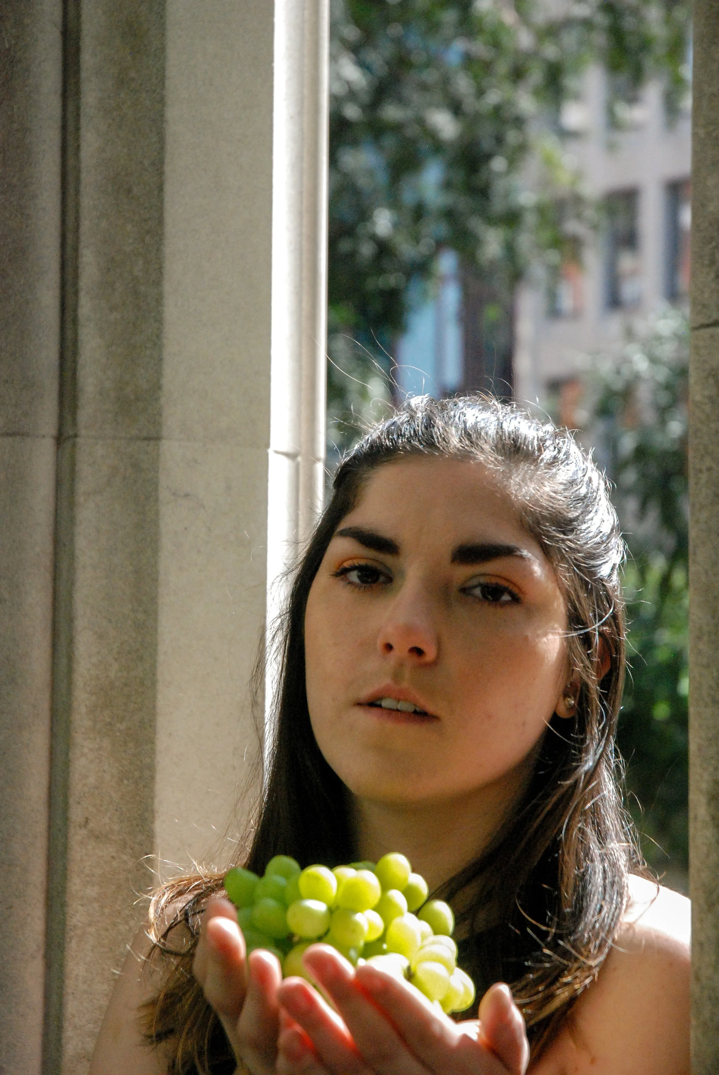 A young woman with long dark hair holding a bunch of green grapes in her hands, standing outside with sunlight on her face and a blurred urban background.