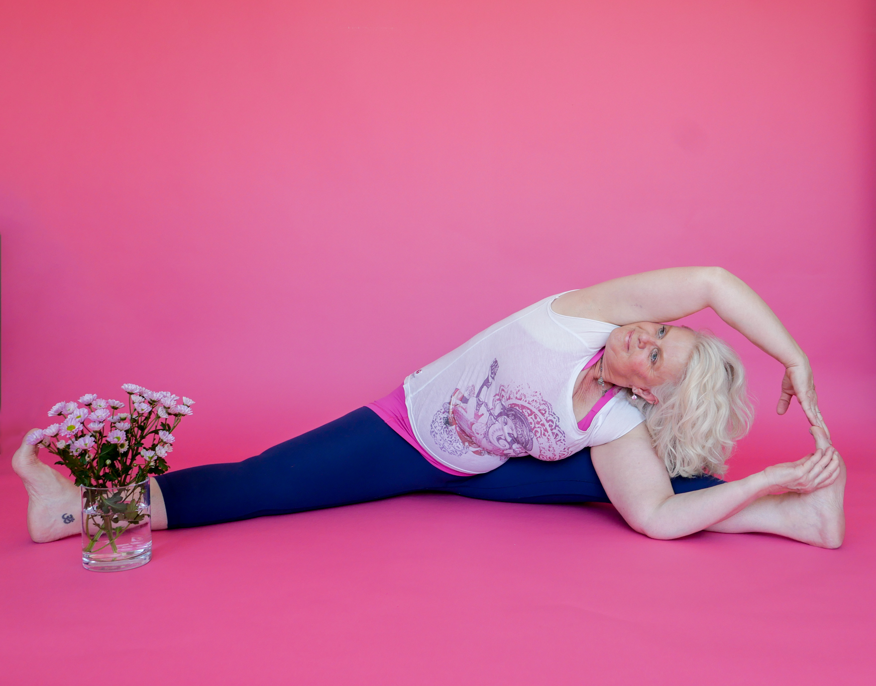 Woman with blonde hair practicing yoga on a pink background, holding a yoga pose while holding a bouquet of pink flowers in a glass vase