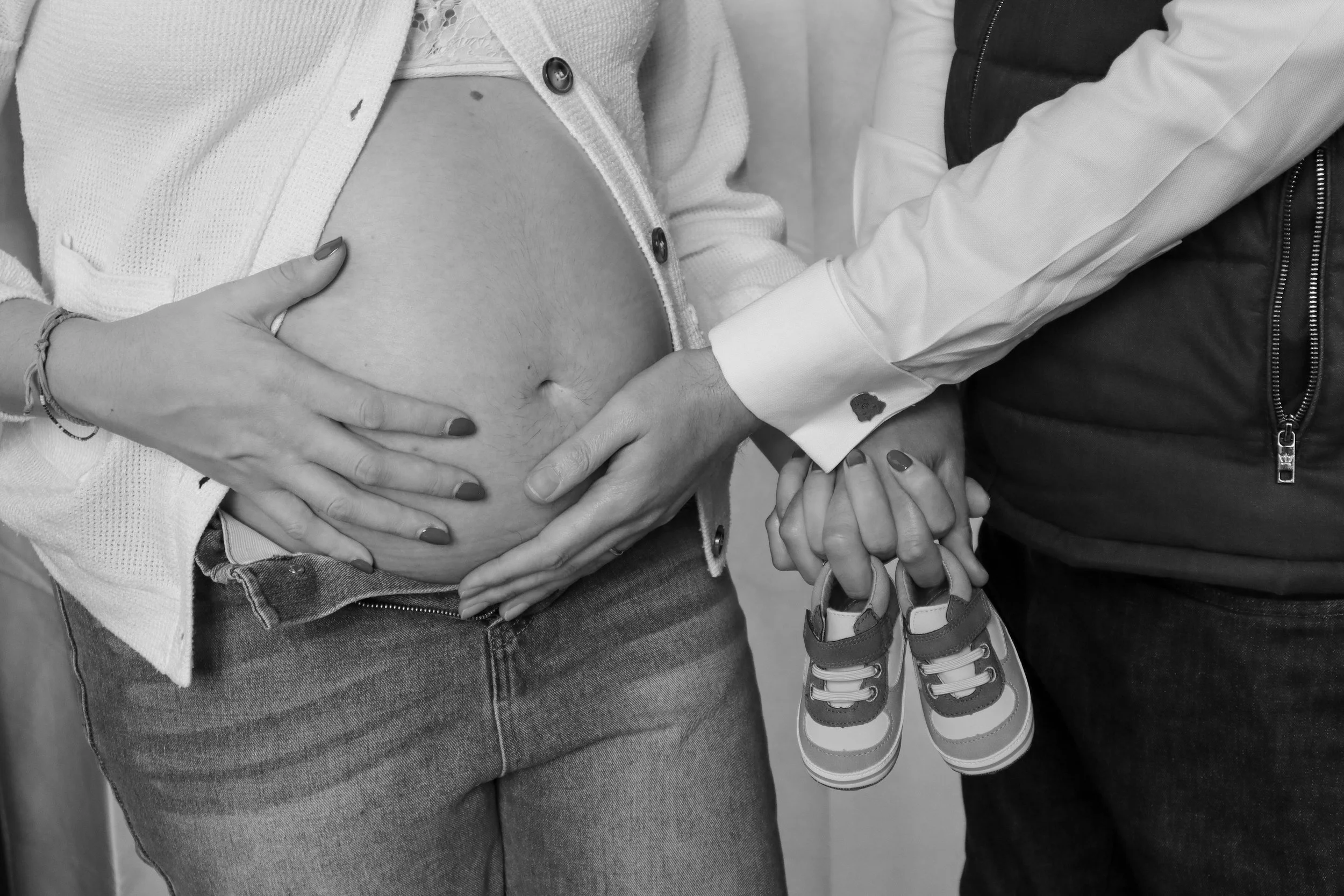 A person wearing a white cardigan and jeans gently holds a pregnant belly, while another person holds a child's hand near a child's feet wearing sneakers.