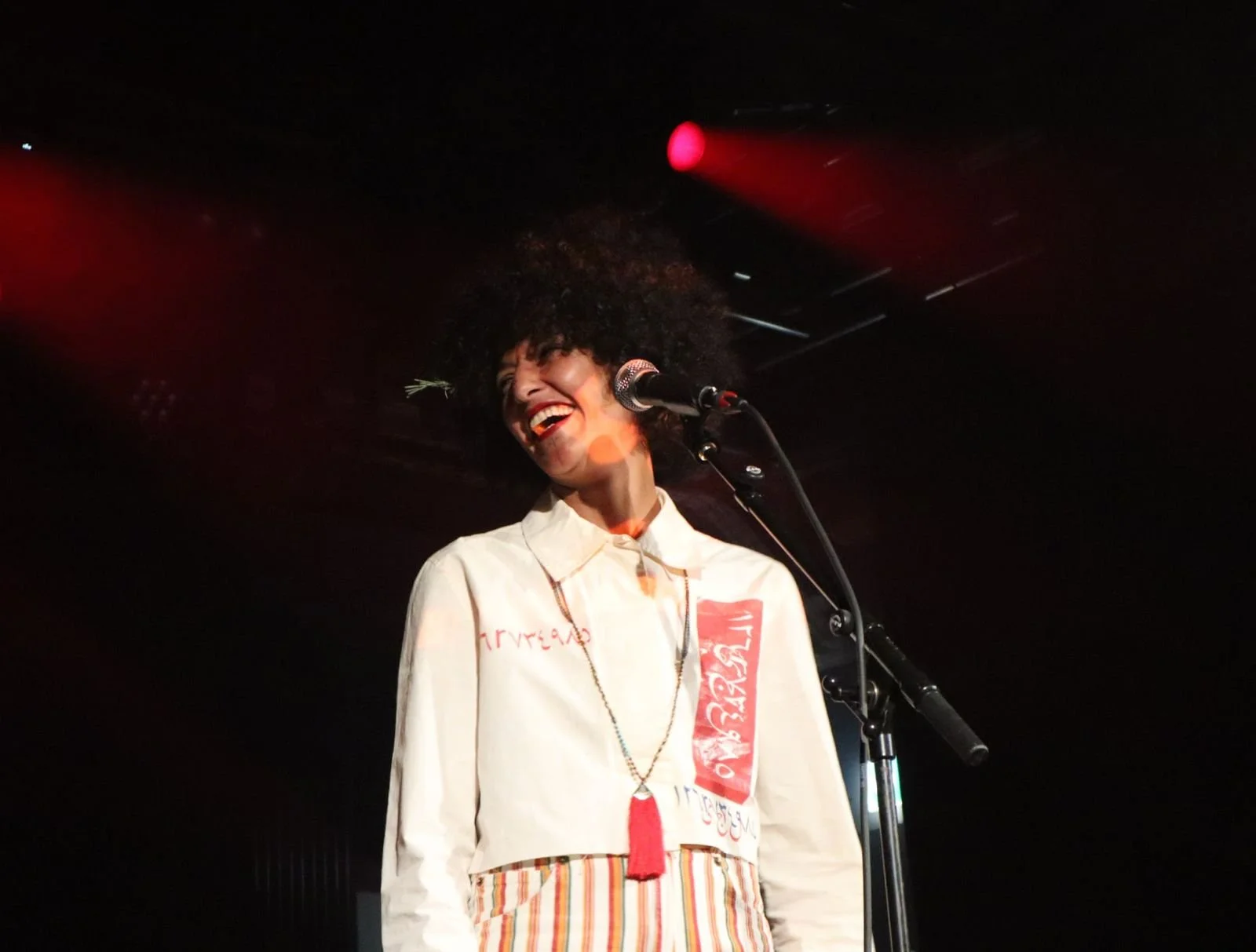A joyful woman with curly hair standing at a microphone on stage, smiling and laughing, wearing a cream-colored jacket with red text and striped pants, with stage lights in the background.
