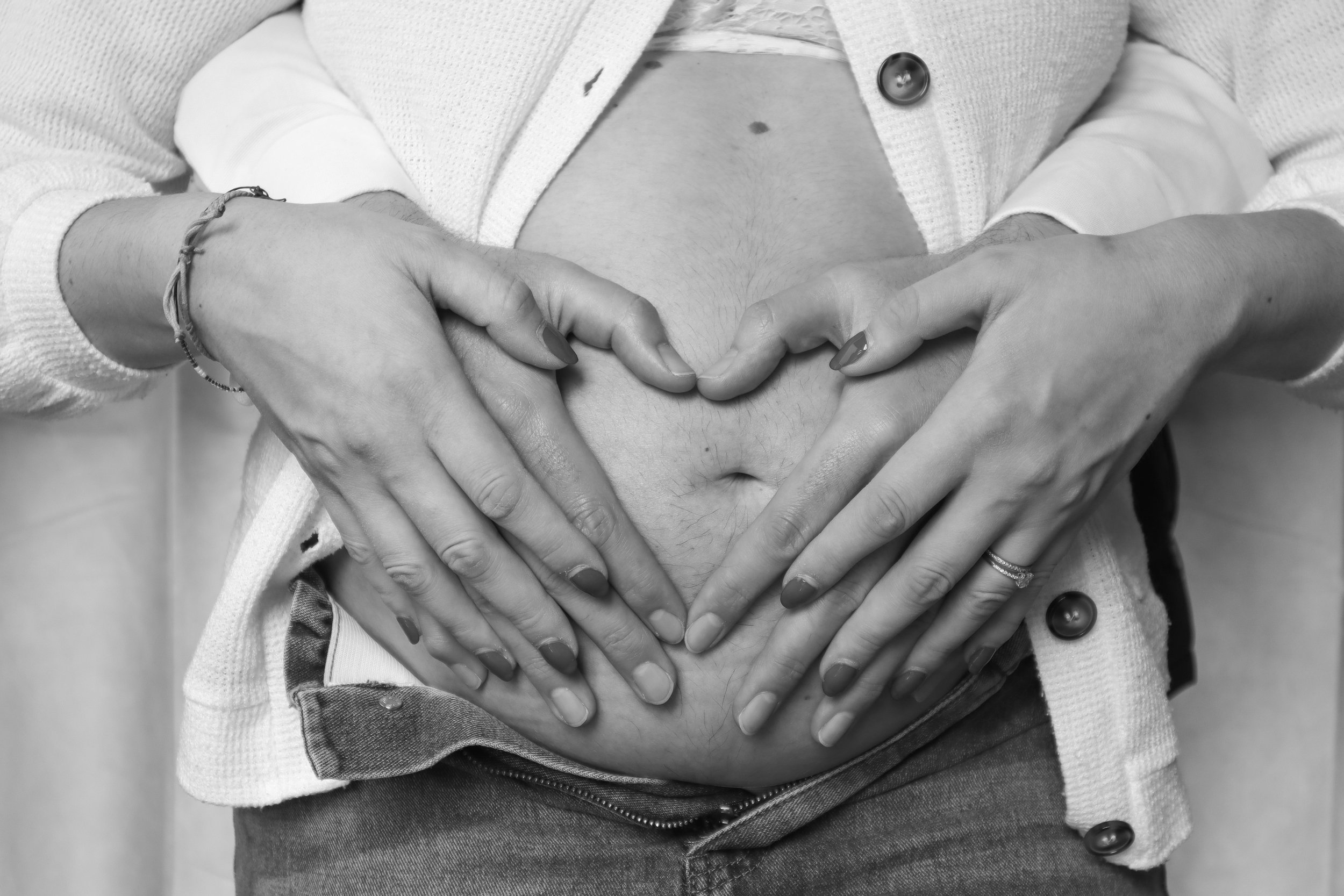 Black and white photo of a person with hands on their stomach in the shape of a heart. The person is wearing a white sweater and has a watch and bracelets on their wrist.