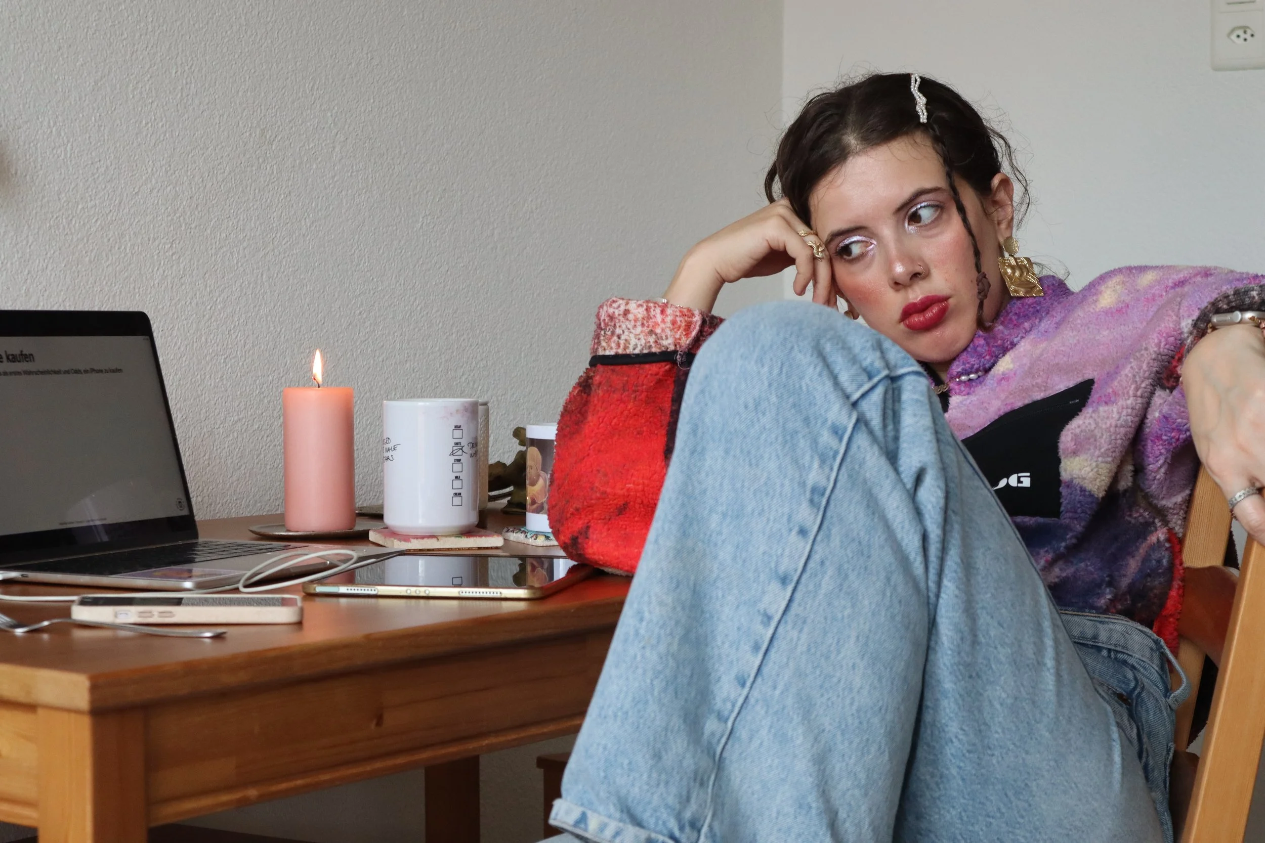 Young woman with dark hair, red lipstick, and jewelry, sitting at a wooden table with her head resting on her hand, surrounded by a laptop, candles, and cups.