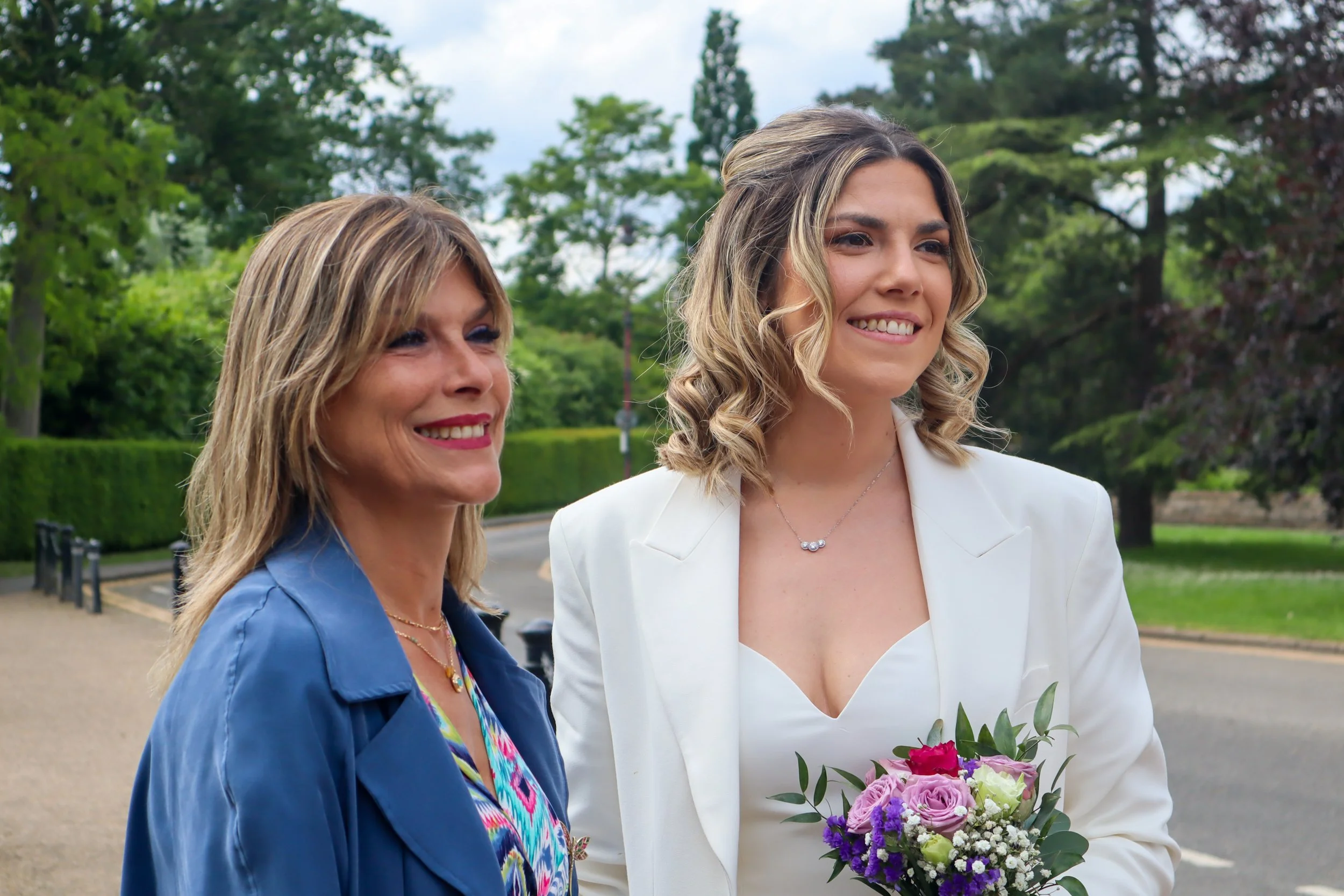 Two women smiling outdoors, one holding a bouquet of pink and purple flowers, surrounded by green trees and a cloudy sky.